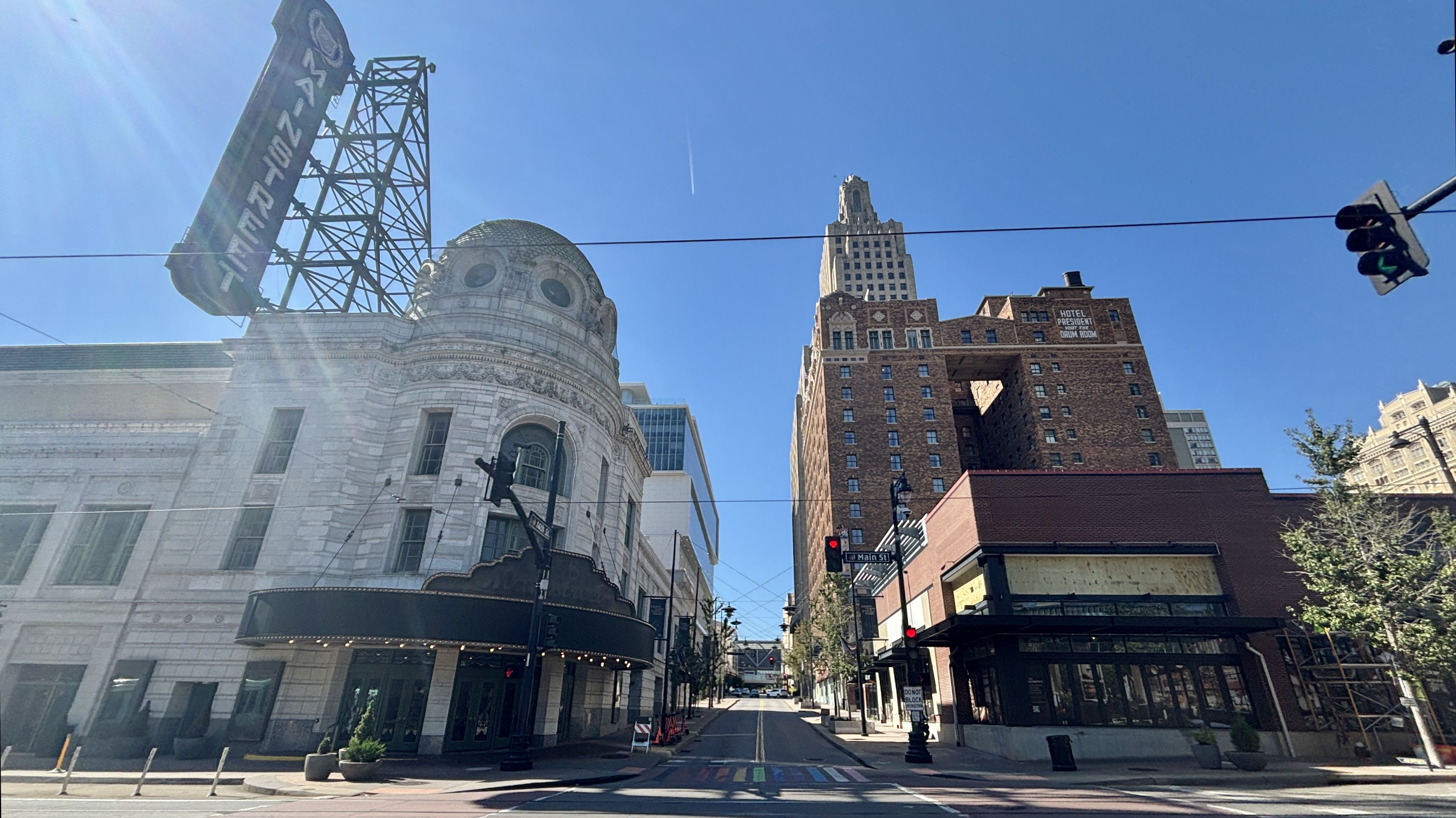 Bright city street view with a white ornate curved building on the left, a tall brick tower on the right, blue sky, sun glare, overhead wires, and a crosswalk at street level.