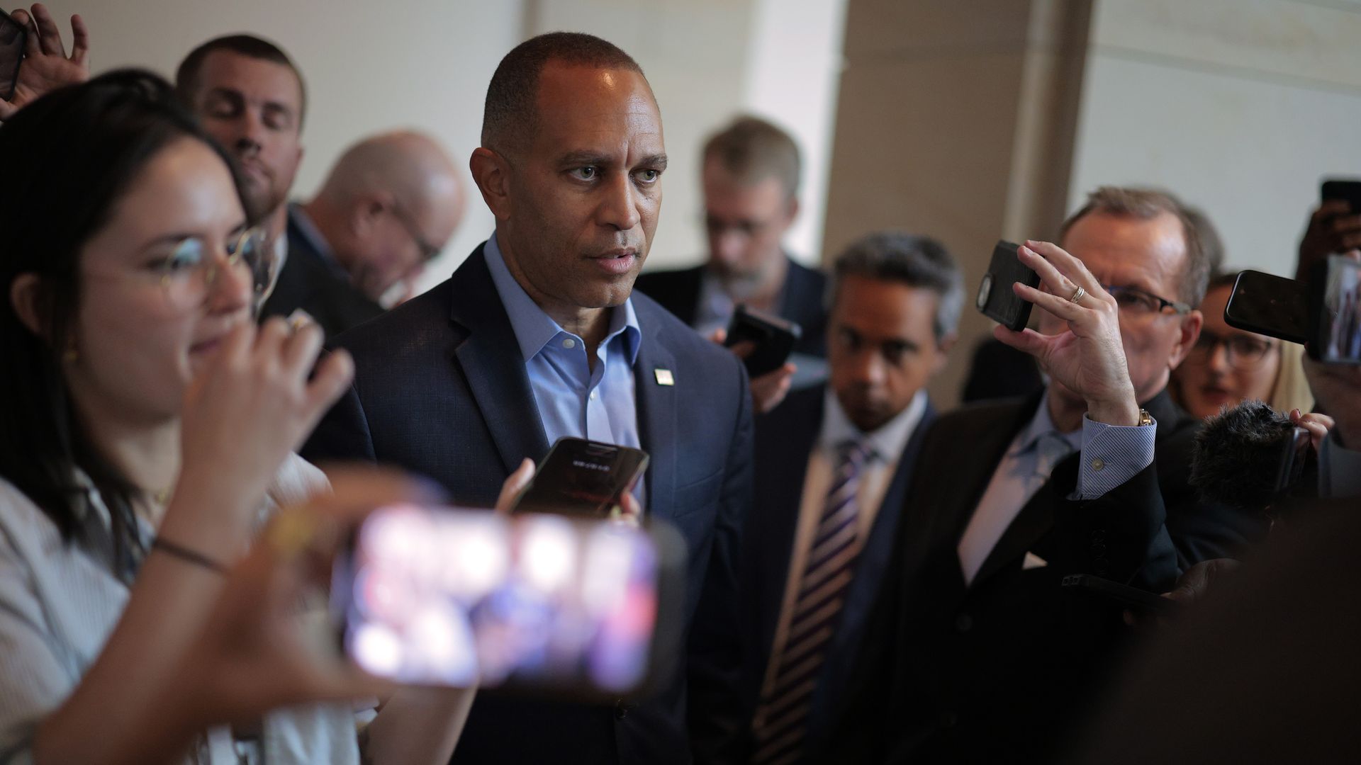 House Minority Leader Hakeem Jeffries, wearing a blue suit, is surrounded by reporters as he walks through a white basement hallway.