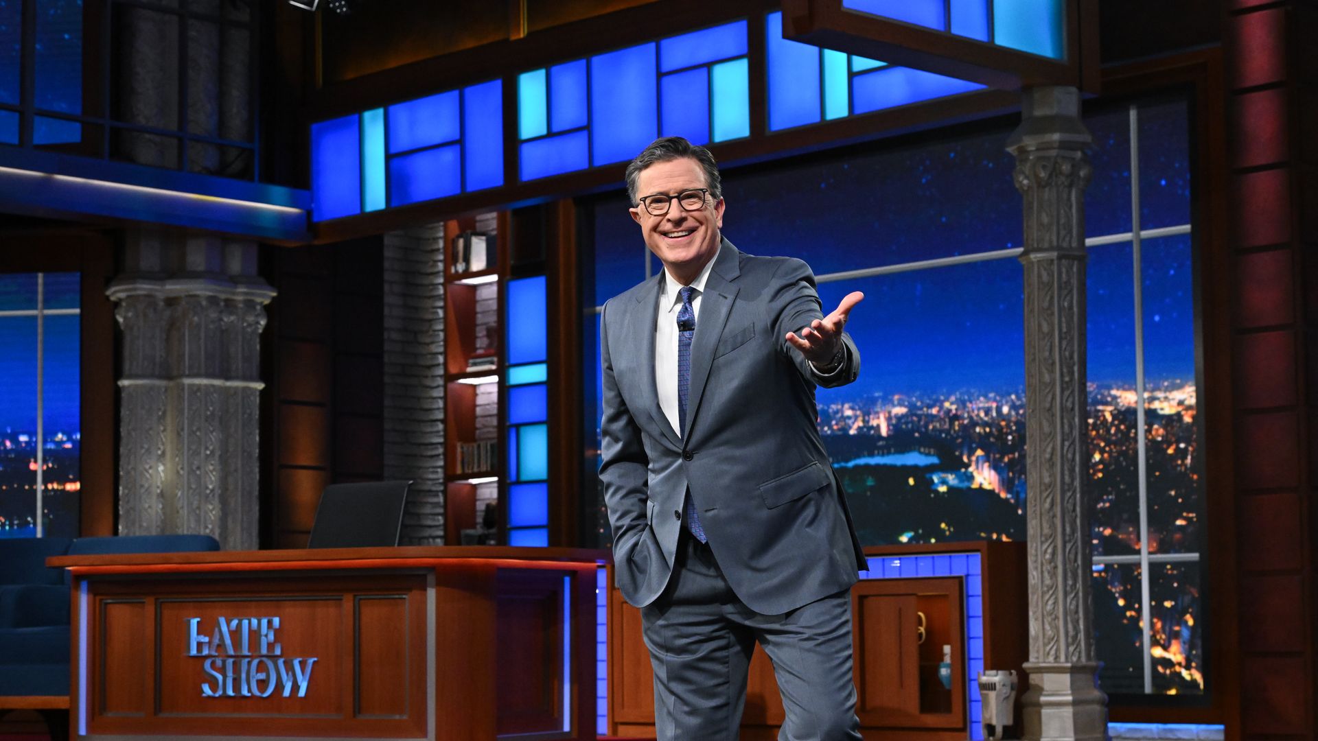 Man in gray suit and glasses smiles and gestures on a blue-lit late night talk show set with a wooden desk labeled "Late Show" against a night cityscape backdrop.
