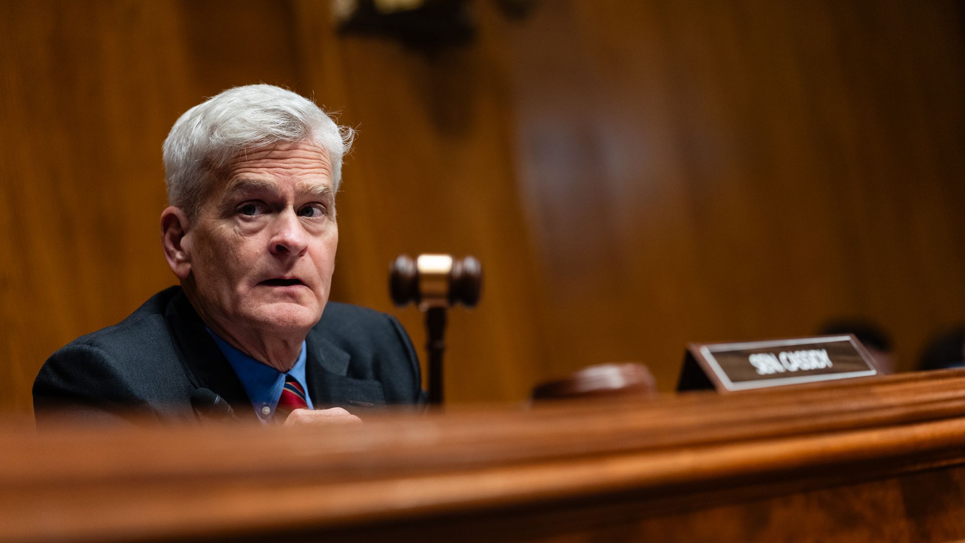 A man with white hair wearing a dark suit jacket holds a gavel as he sits in a Capitol Hill confirmation hearing behind a small sign that says "Sen. Cassidy."