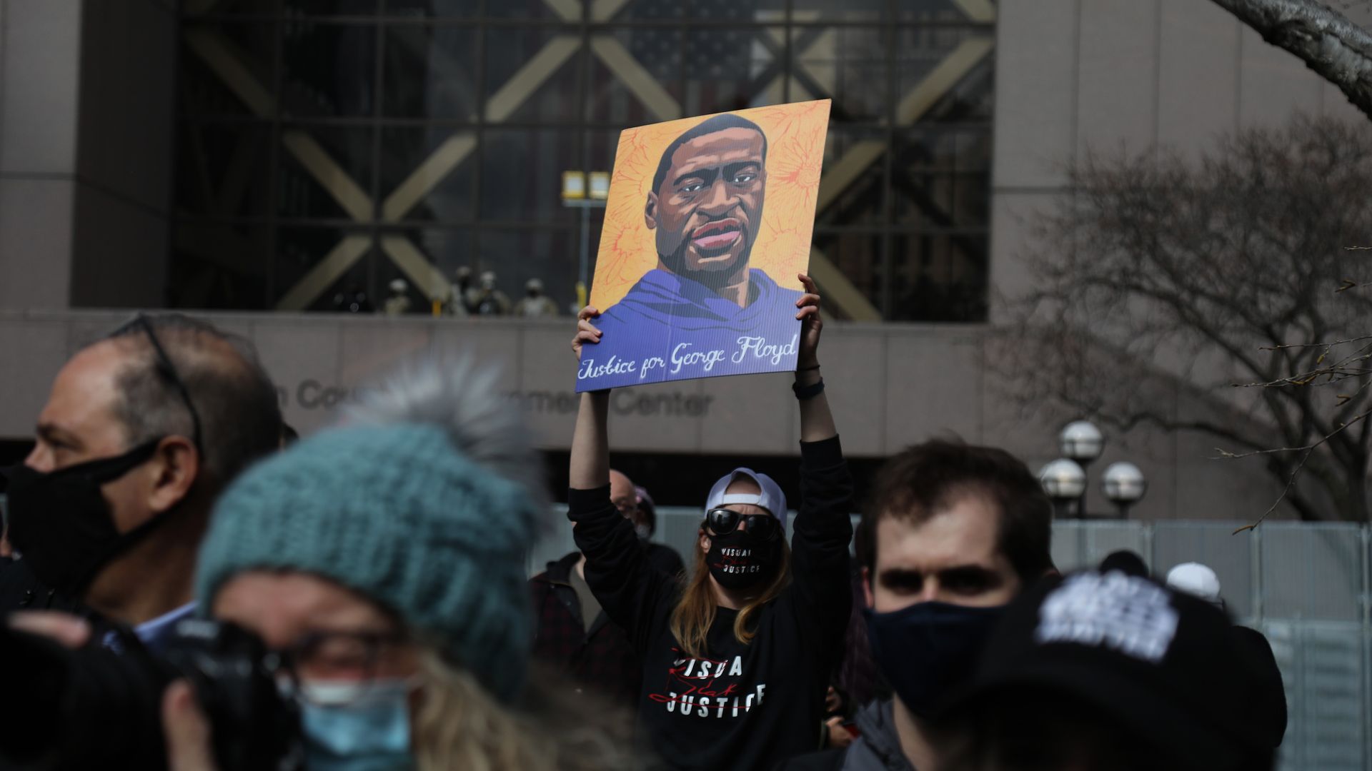 Photo of a masked protester holding up a sign bearing a painted representation of George Floyd