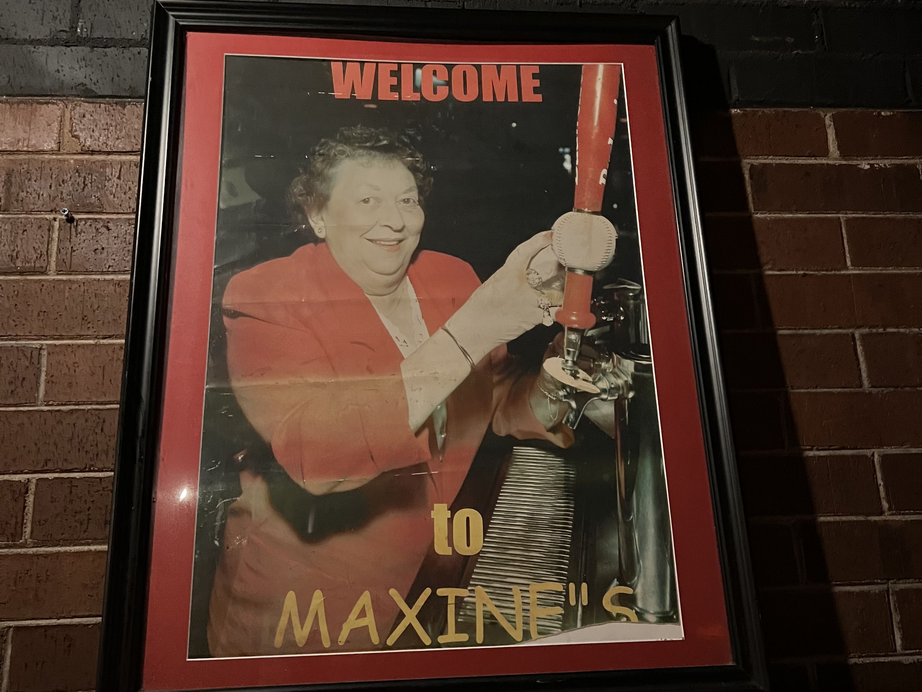 framed photo on a brick wall of an older woman in a red sport coat pouring beer from taps