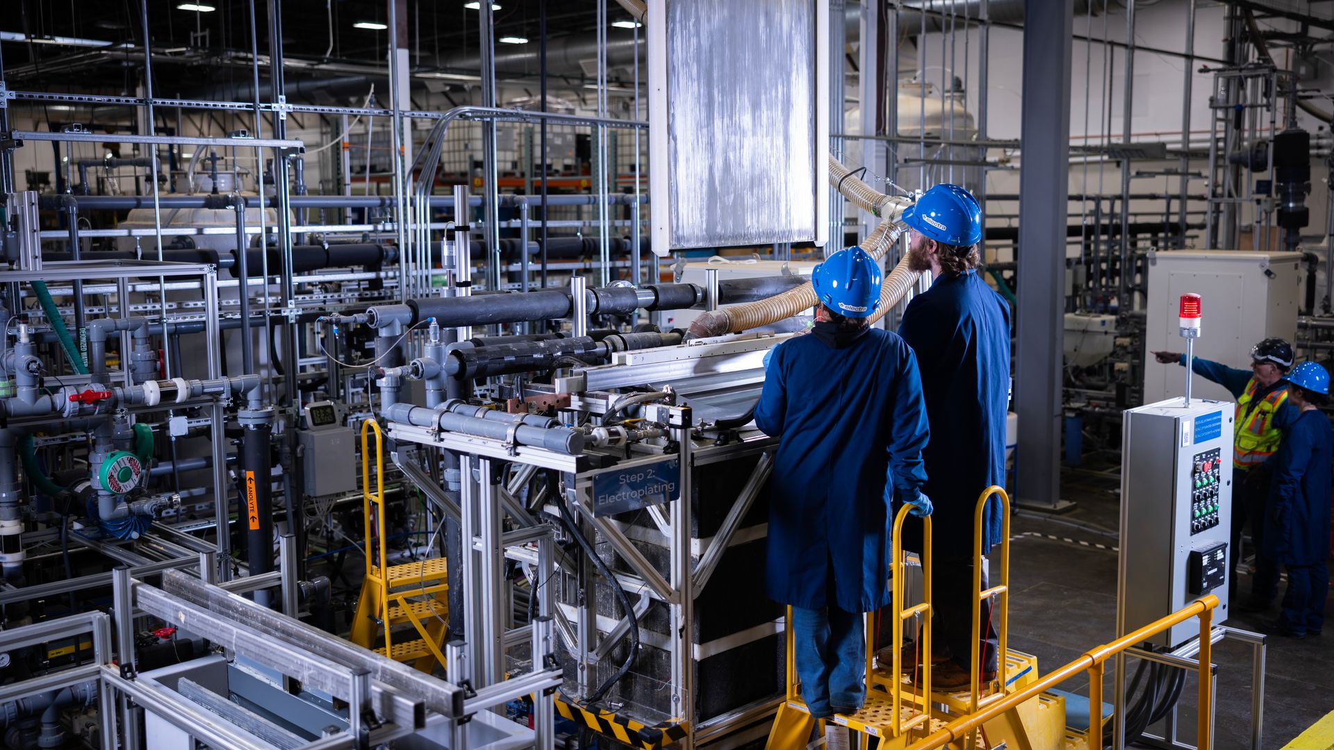 Two workers in blue coats and helmets operate machinery labeled Step 2 Electroplating in a factory, while two others in safety vests and helmets stand nearby amid pipes and equipment.