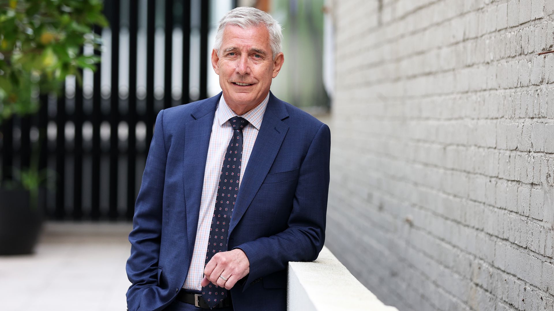 A person with gray hair wearing a navy blue suit, patterned tie, and checked shirt, standing outdoors by a white ledge and a brick wall, smiling at the camera.