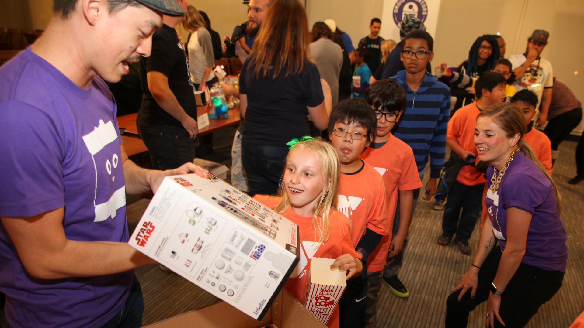 In this image, a man hands out toy building and code kits to a line of children. The first child in line is a young blonde girl. 