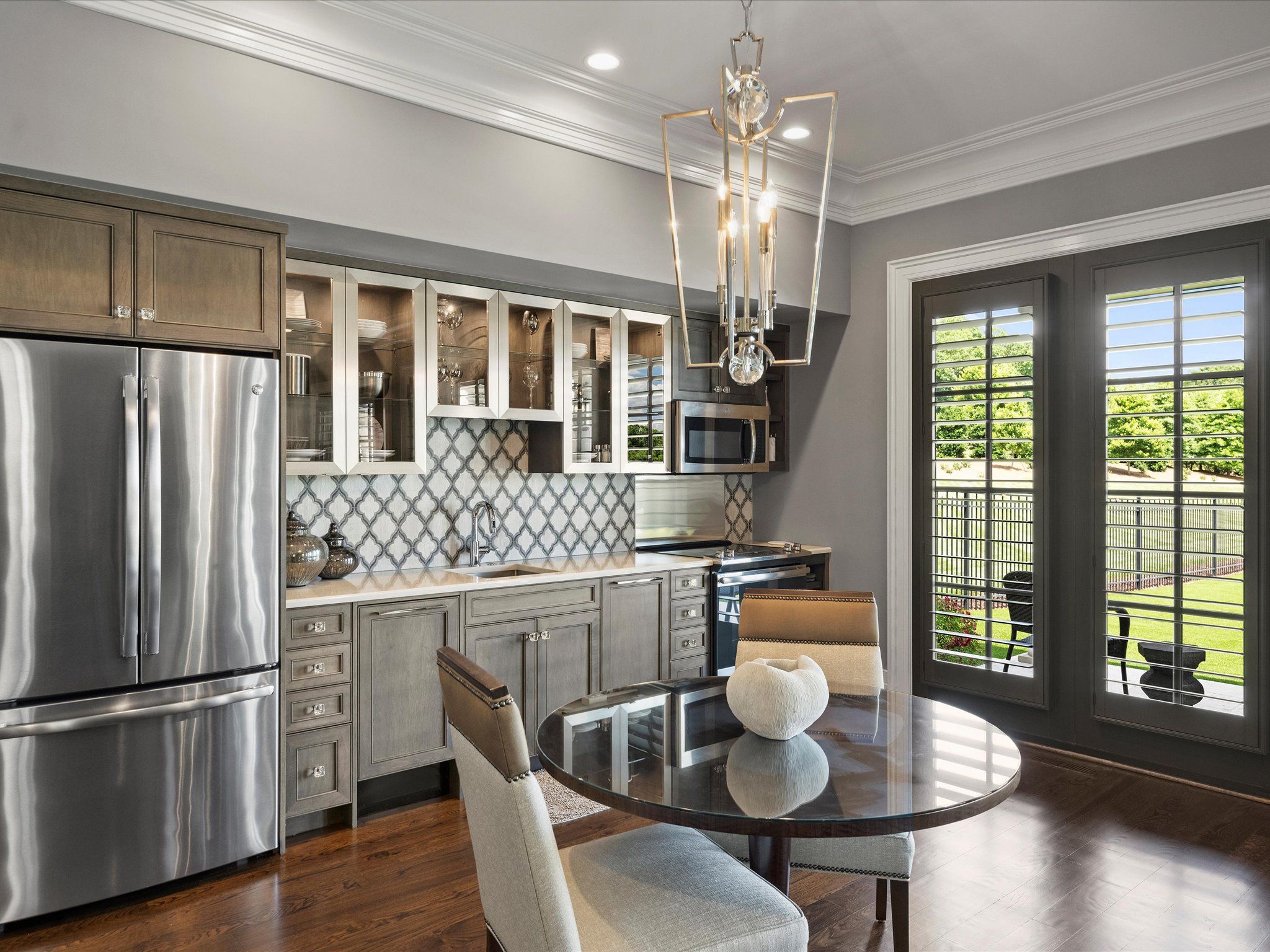 Modern kitchen with gray cabinetry, stainless steel appliances, patterned backsplash, glass dining table, cushioned chairs, and large windows opening to a green backyard.