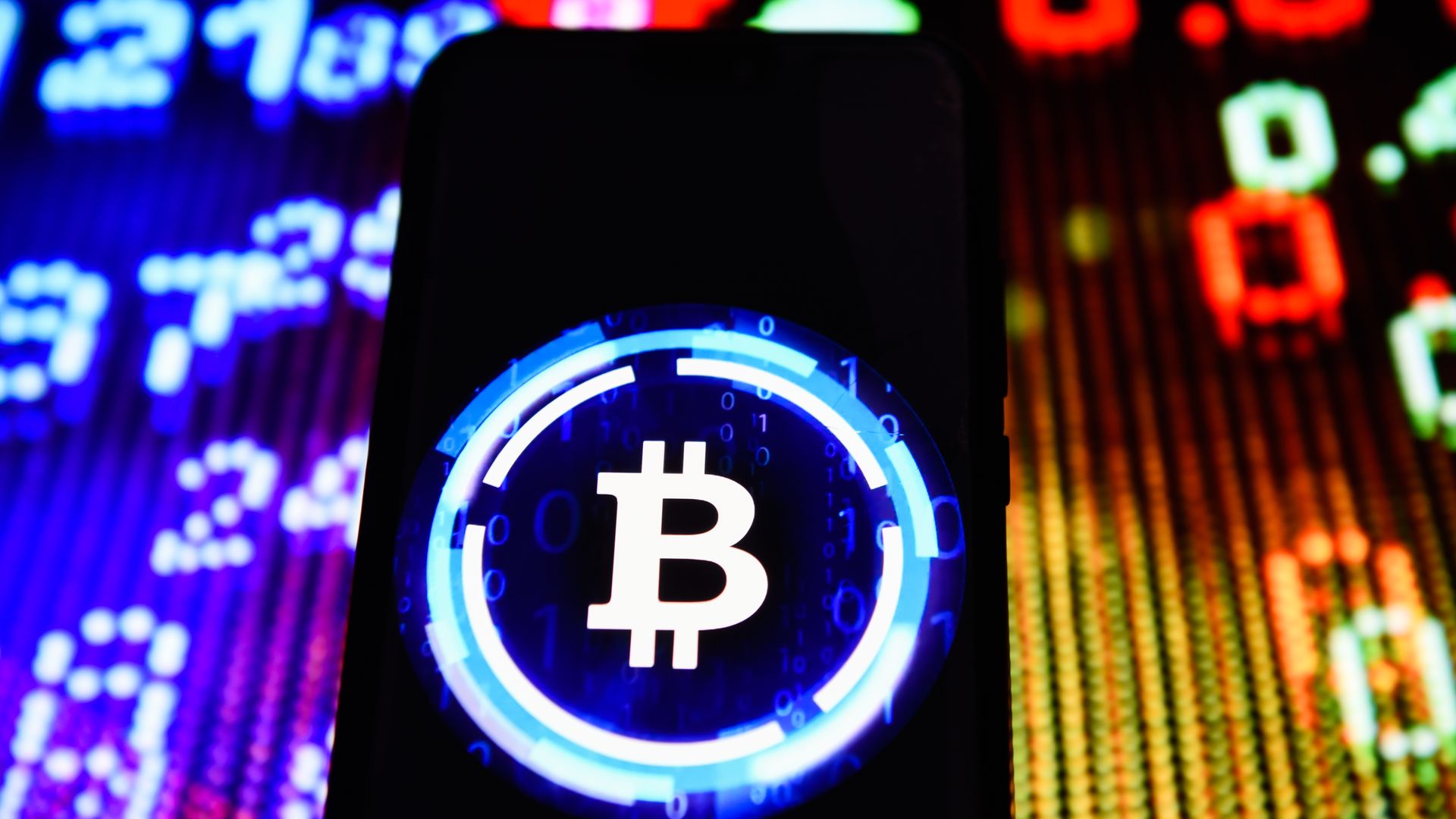 In this image, the neon blue and white Bitcoin logo is highlighted on a black phone screen in front of a bright colored background of numbers.
