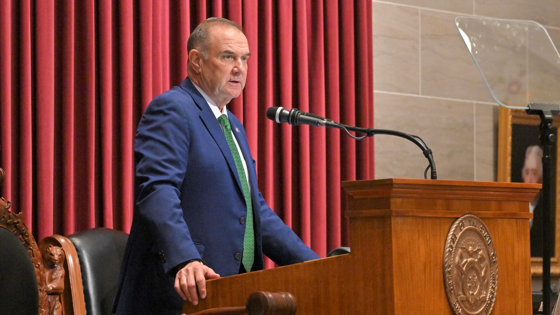 Missouri Gov. Mike Kehoe in a blue suit with a green tie giving his state of the state address at a wooden podium with a microphone and red curtains in the background.