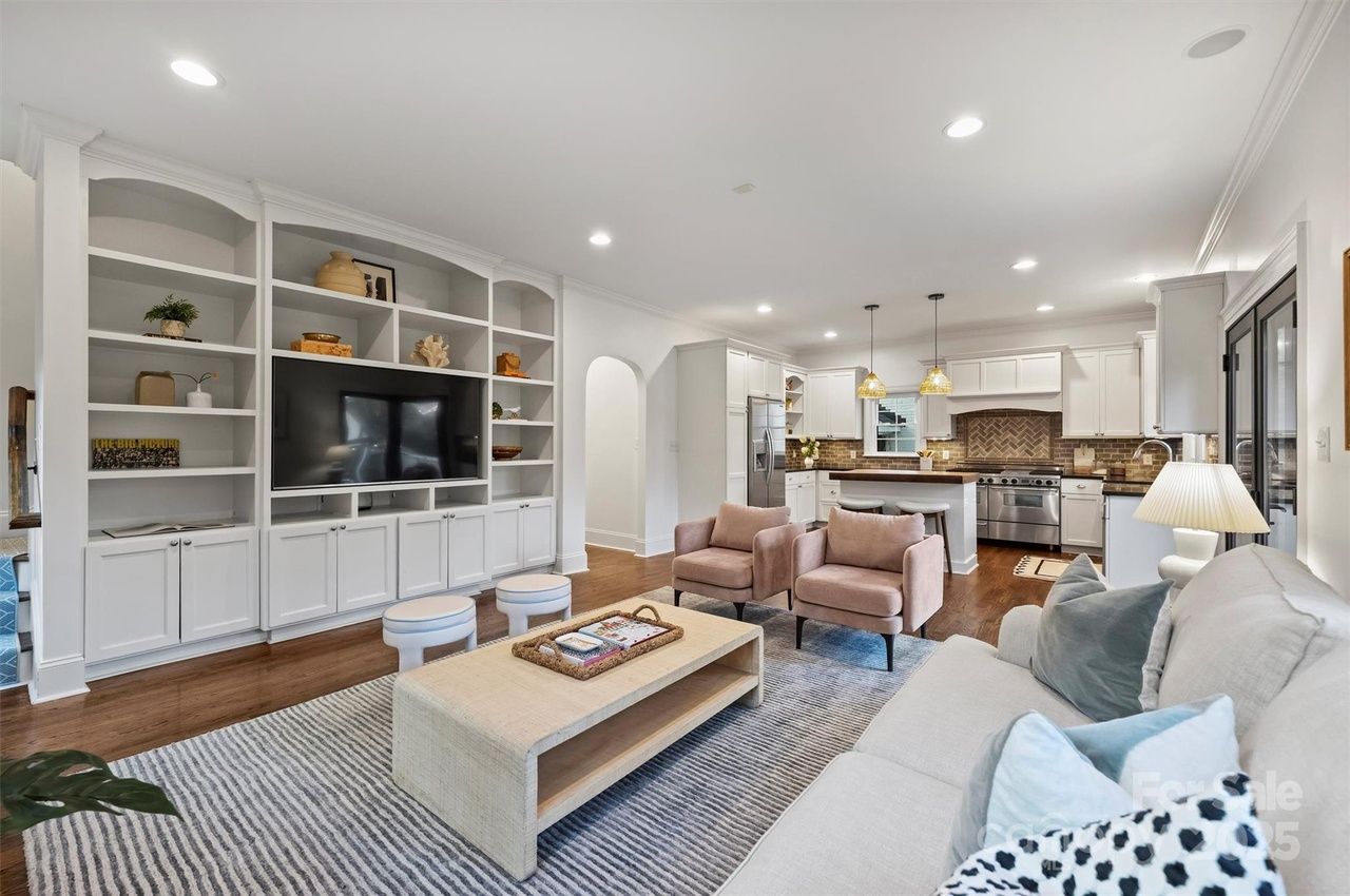 Bright open living room and kitchen with white cabinetry, hardwood floors, beige sofa, two pink armchairs, large TV, striped rug, and pendant lights over kitchen island.