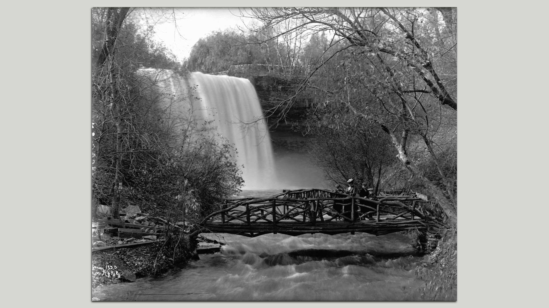 Black-and-white image of a wide waterfall over a rocky ledge, with a wooden railing bridge in the foreground and people sitting along it among bare trees.