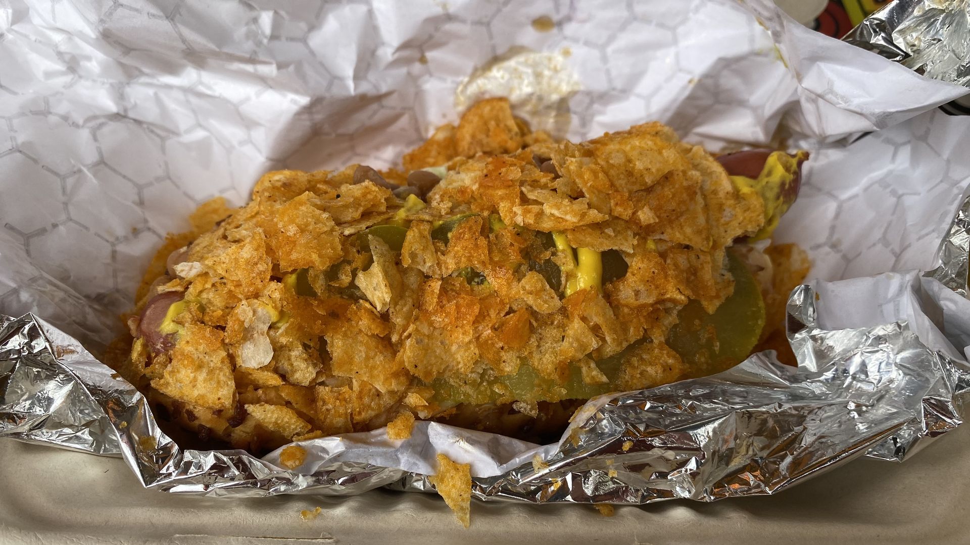 A hot dog on a foil wrapper covered in mustard, a pickle and crumbled potato chips.