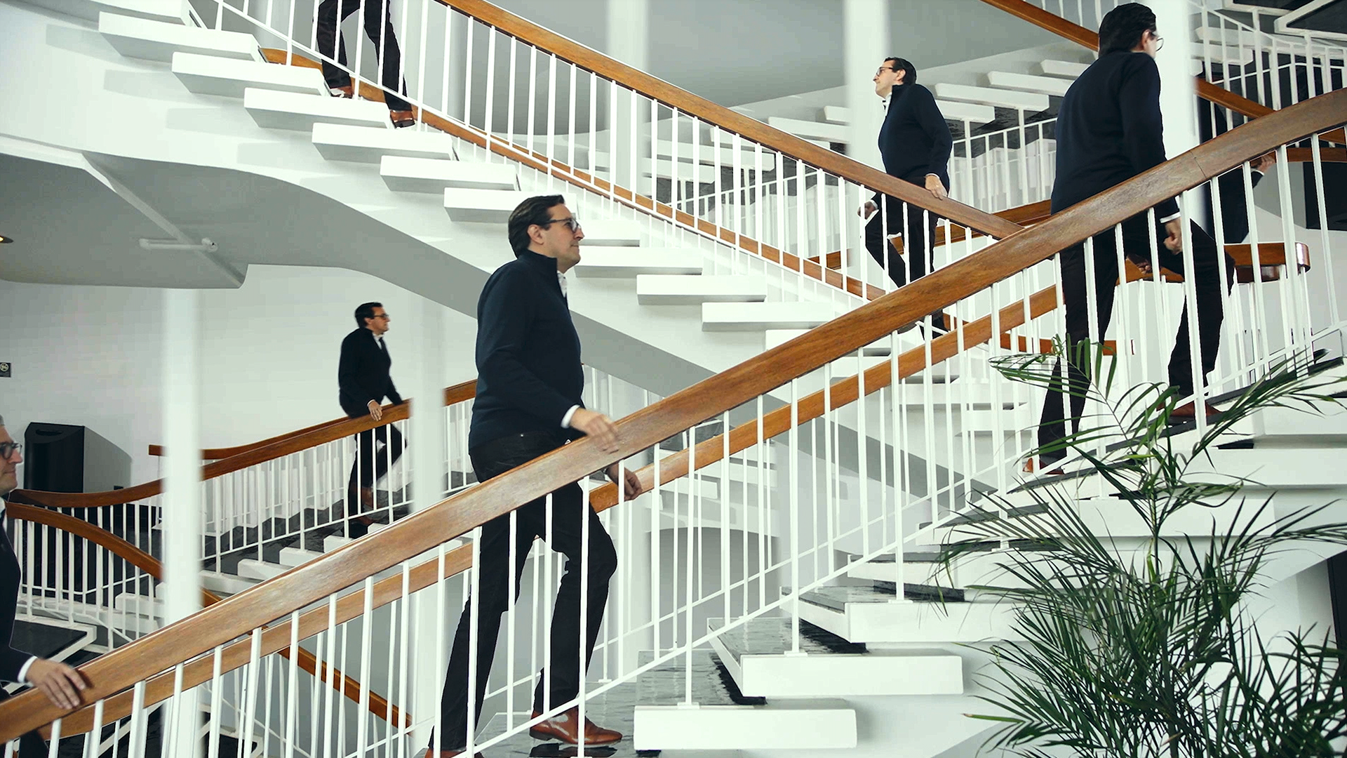 Multiple images of a man in black clothing and brown shoes walking up white stairs with wooden handrails inside a modern building with green plants.