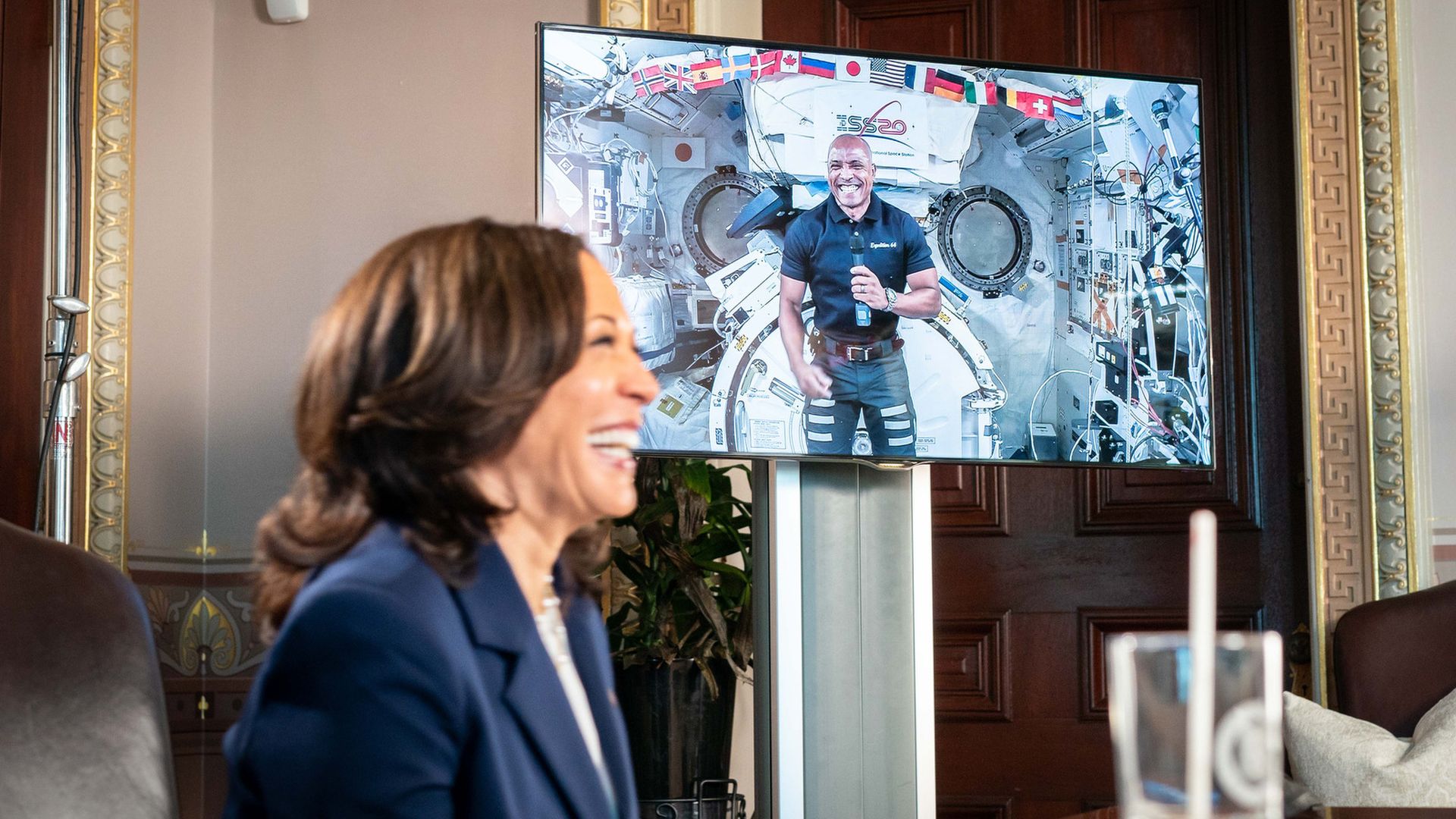 Kamala Harris smiles in the foreground while speaking with astronaut Victor Glover in space, pictured on a TV to the side of her.