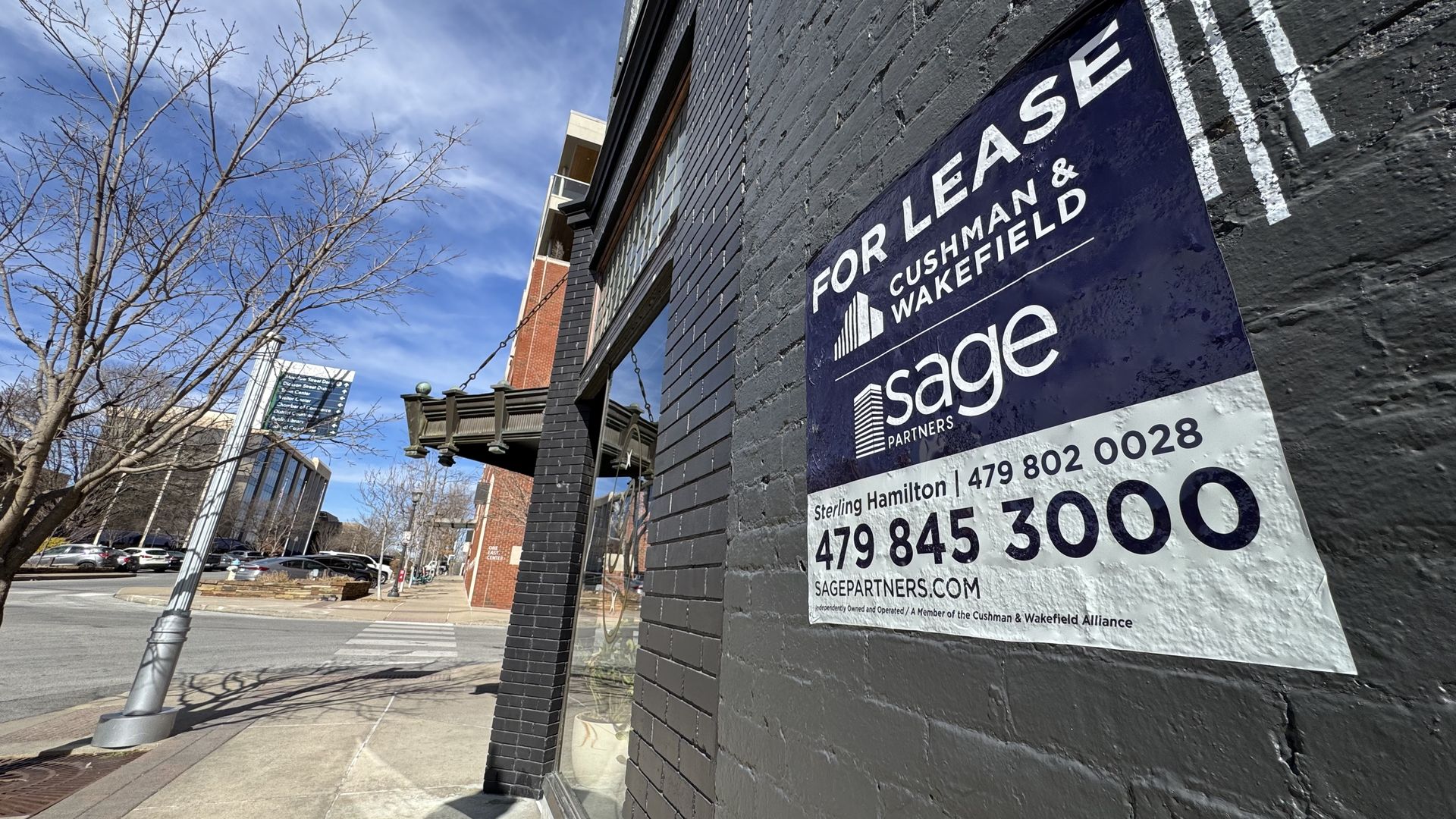 A "For Lease" sign is mounted on a downtown brick storefront, with a sidewalk, street, and nearby buildings under a blue sky.