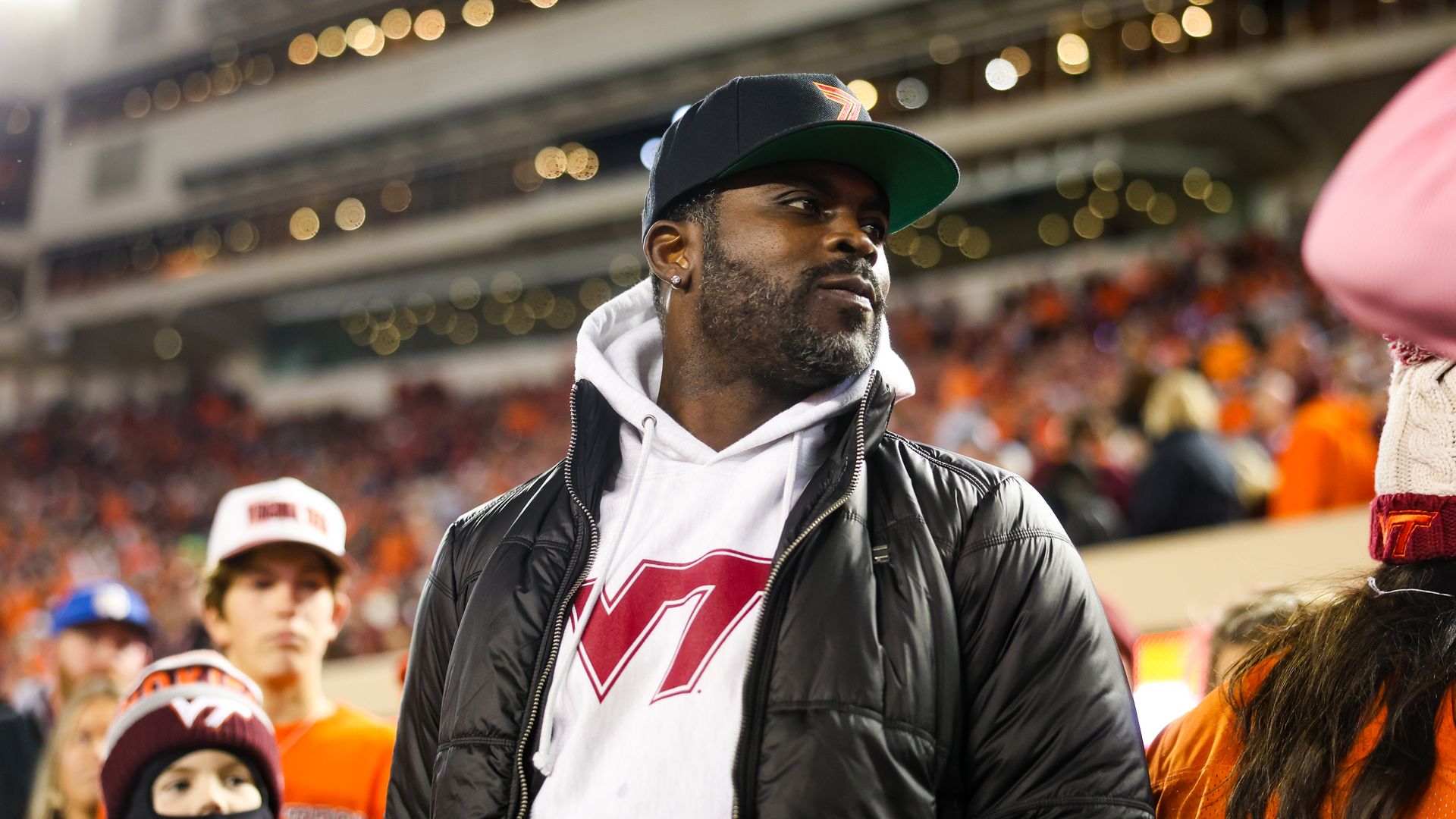 A man stares off to the right hand side while wearing a white VT hoodie, black jacket and a black hat