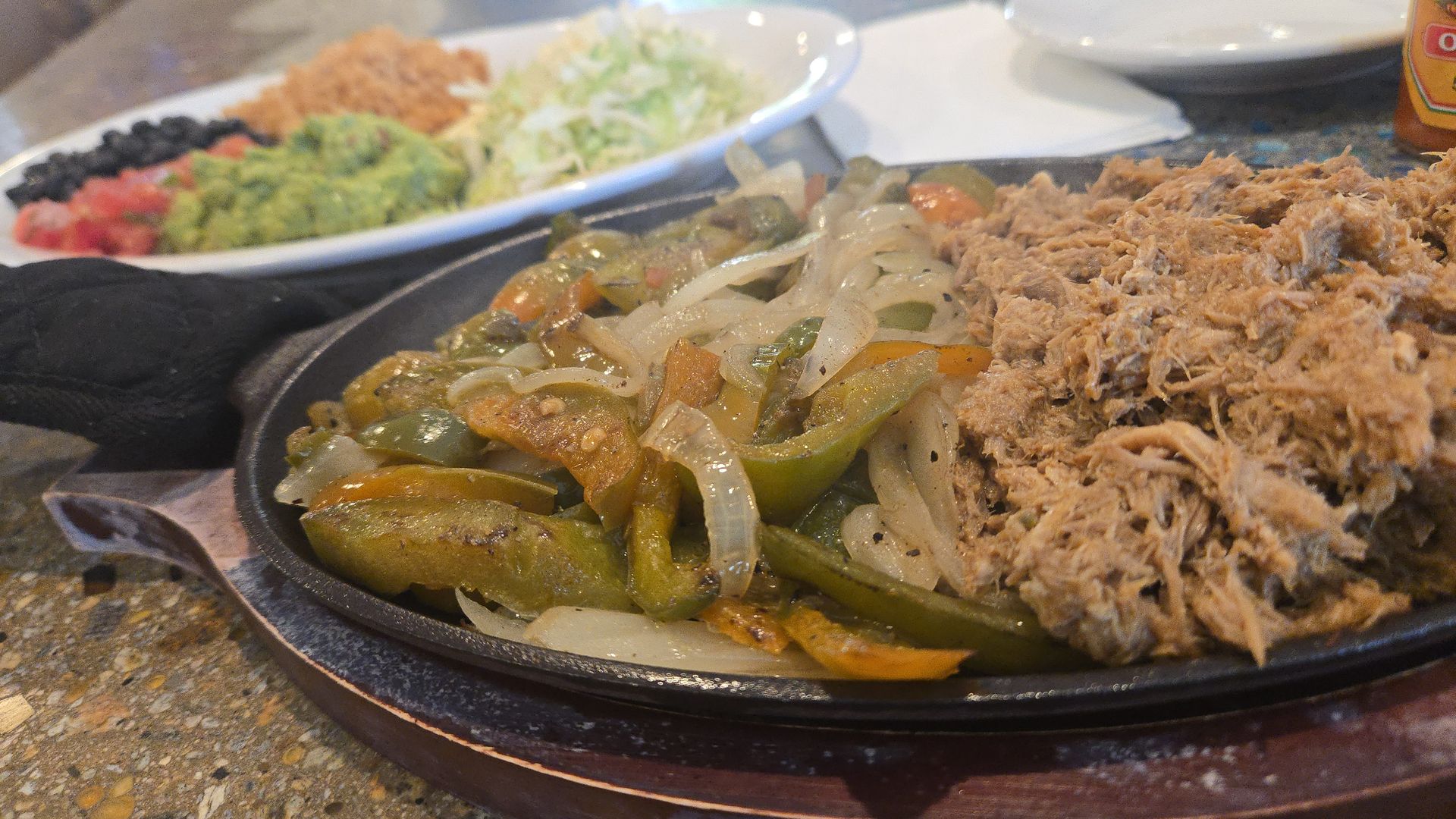 Sizzling fajita plate with sautéed onions and bell peppers in the foreground and shredded pork on the right; a background plate holds guacamole, pico de gallo, lettuce.