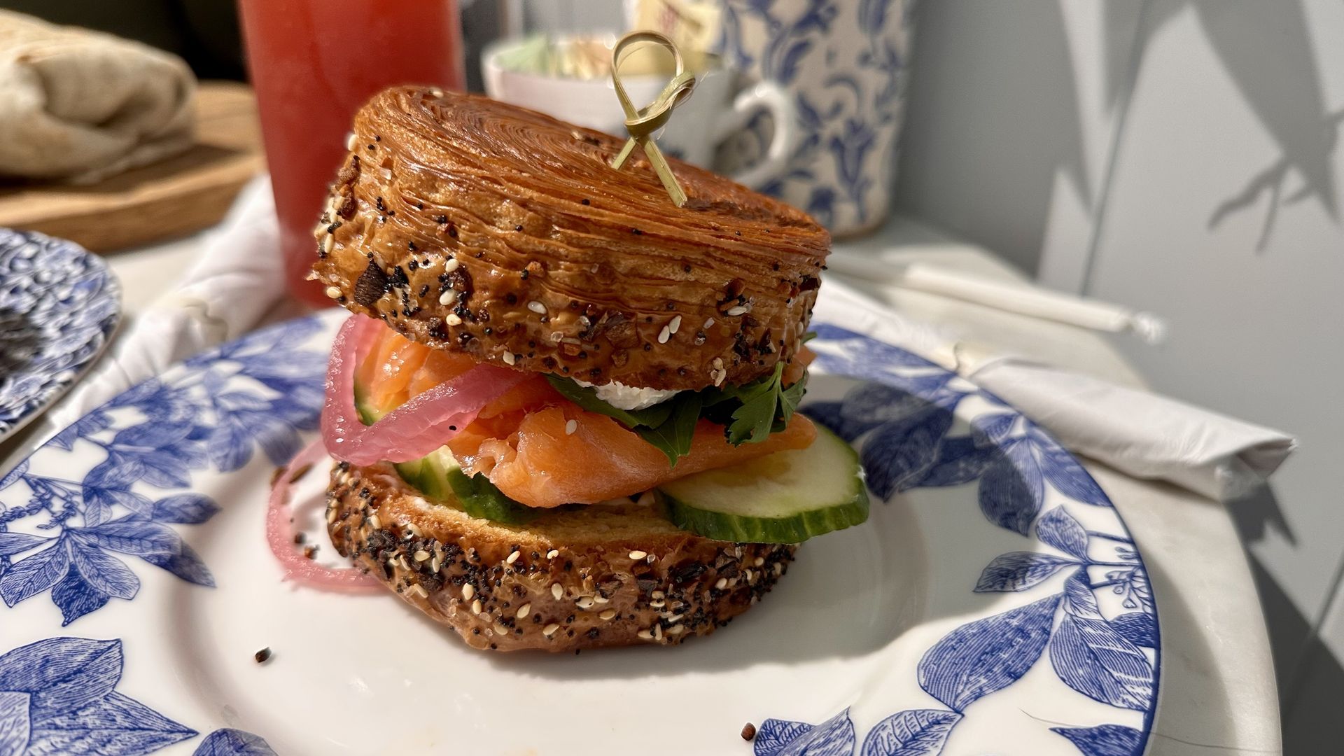 Smoked salmon sandwich with cucumber, pickled red onion, greens, and cream cheese on an everything croissant, served on a white plate with blue leaf pattern.