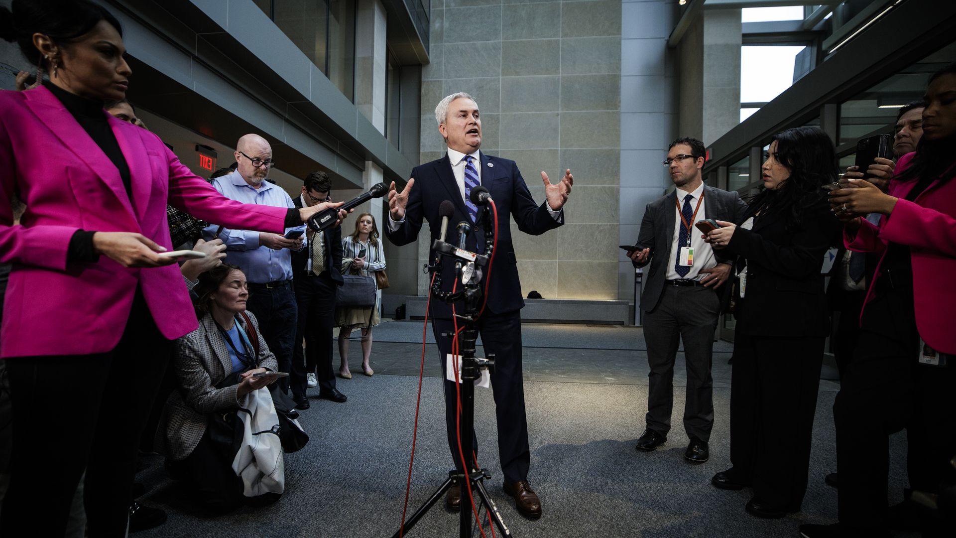 Rep. James Comer, wearing a blue suit, white shirt and blue striped tie, speaks to reporters while standing at a microphone in a gray, metallic building.
