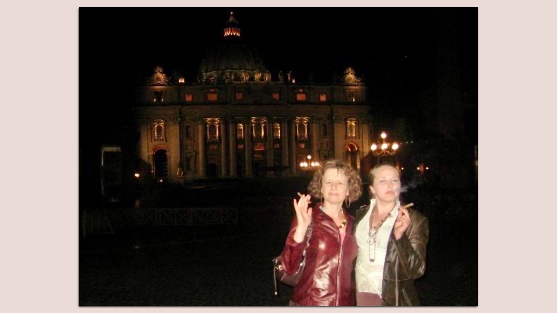 Two women in jackets pose and smoke in the foreground at night, in front of a large, lit historic building with a central dome and tall columns.