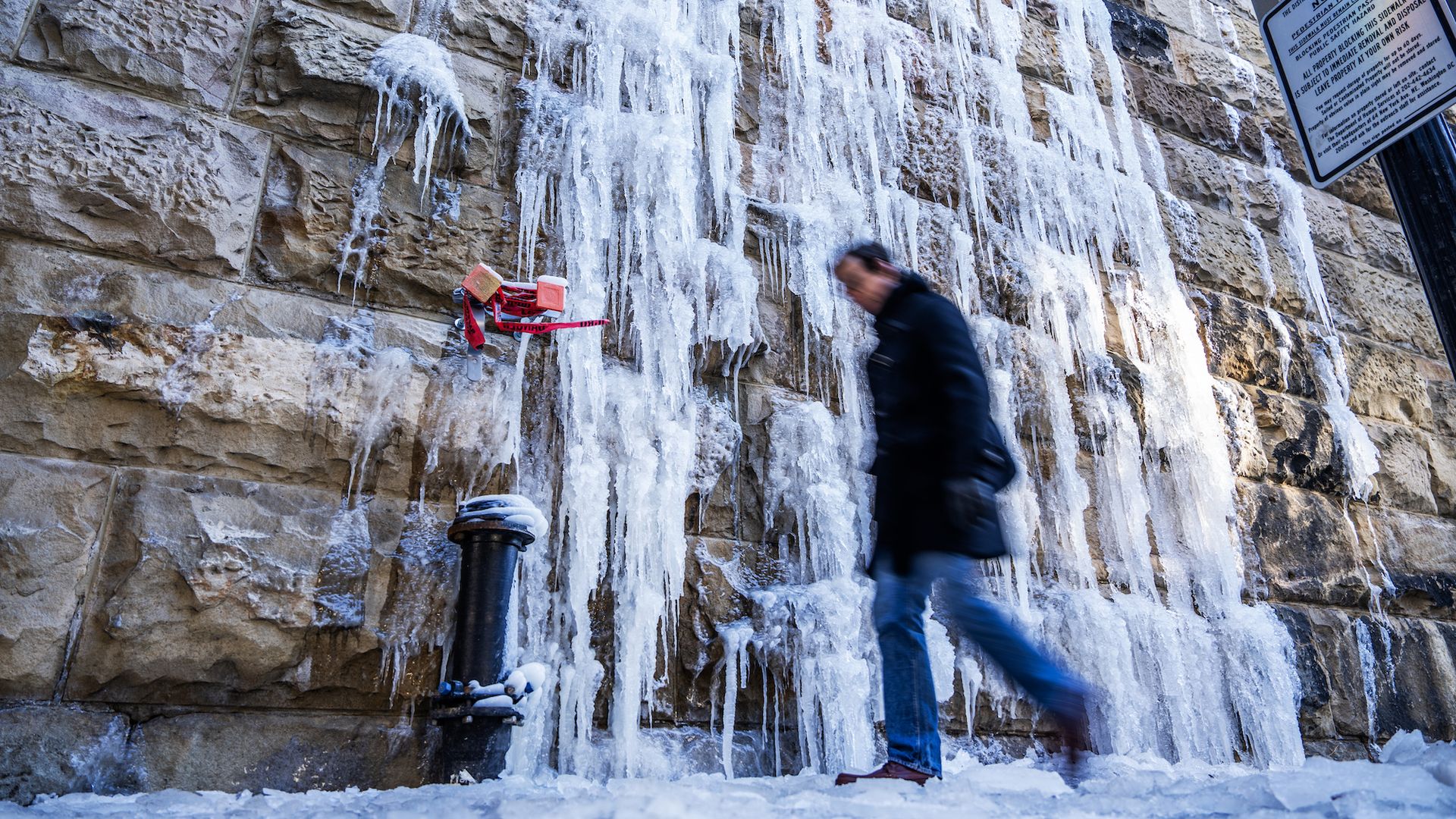 Icicles hanging from a stone wall at Union Station over a black pipe covered in snow, with a person in a coat and gloves walking by. A signpost with a bike yield sign is visible.