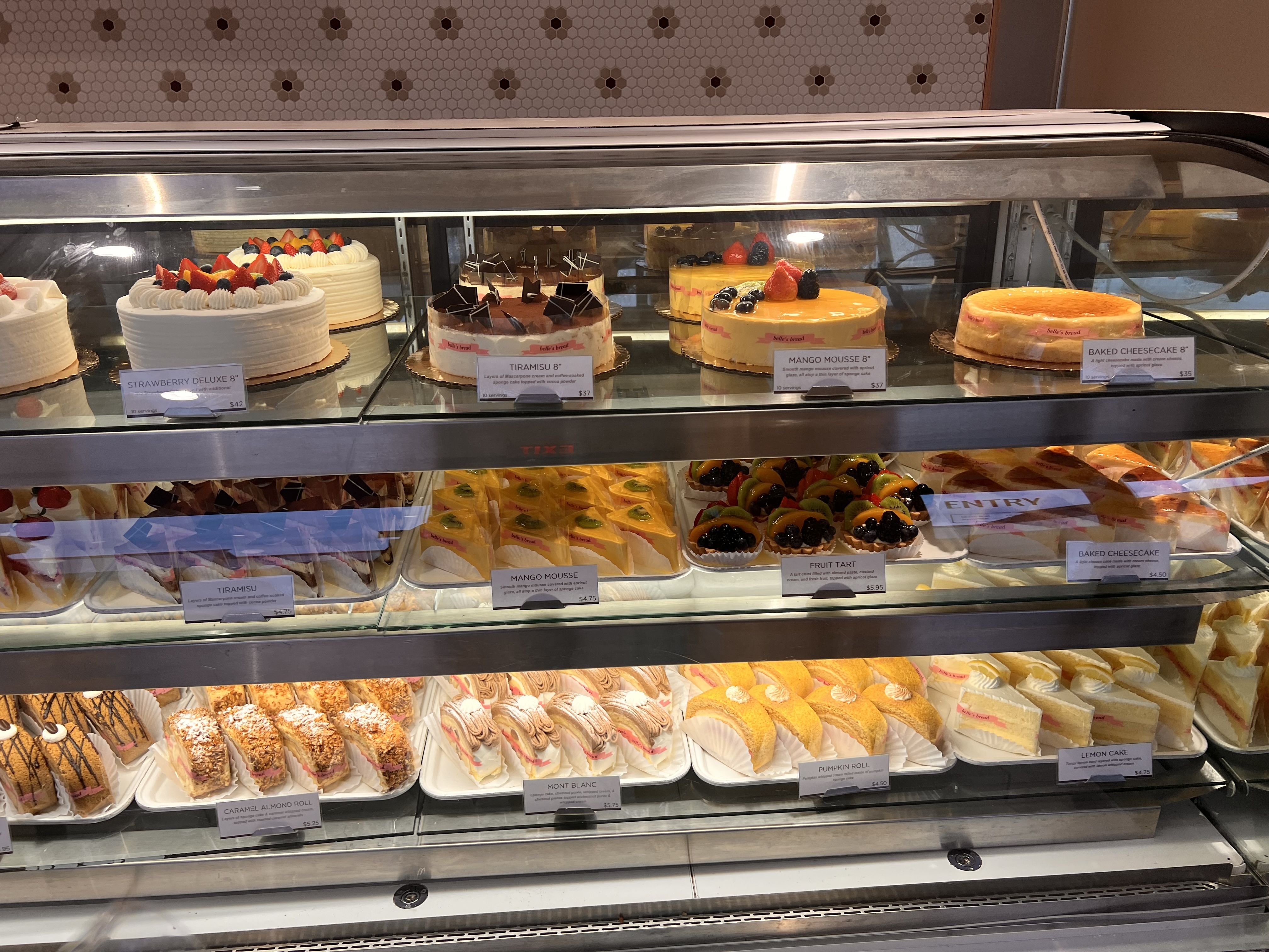 Display case with various cakes and pastries including strawberry deluxe, tiramisu, mango mousse, baked cheesecake, fruit tarts, caramel almond rolls, mont blanc, pumpkin roll, and lemon cake.