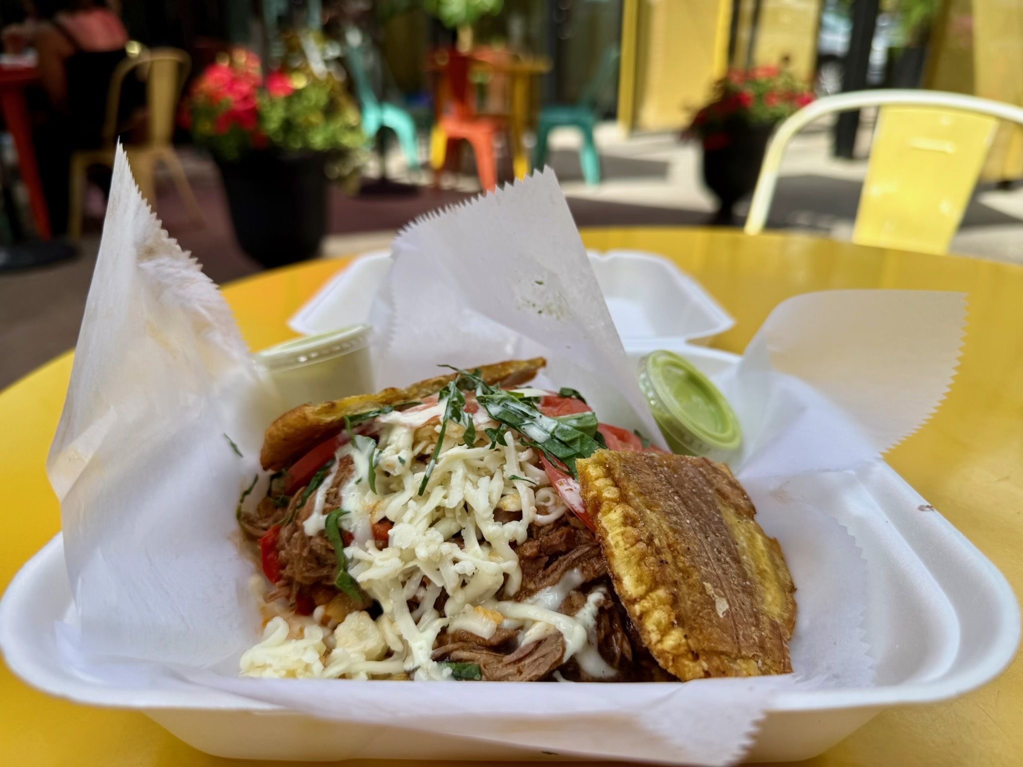 Close-up of a takeout meal on a yellow cafe table. White tray with paper wrapping holds shredded cheese, herbs, tomatoes, and sliced meat; a lime wedge rests nearby, blurred chairs in background.