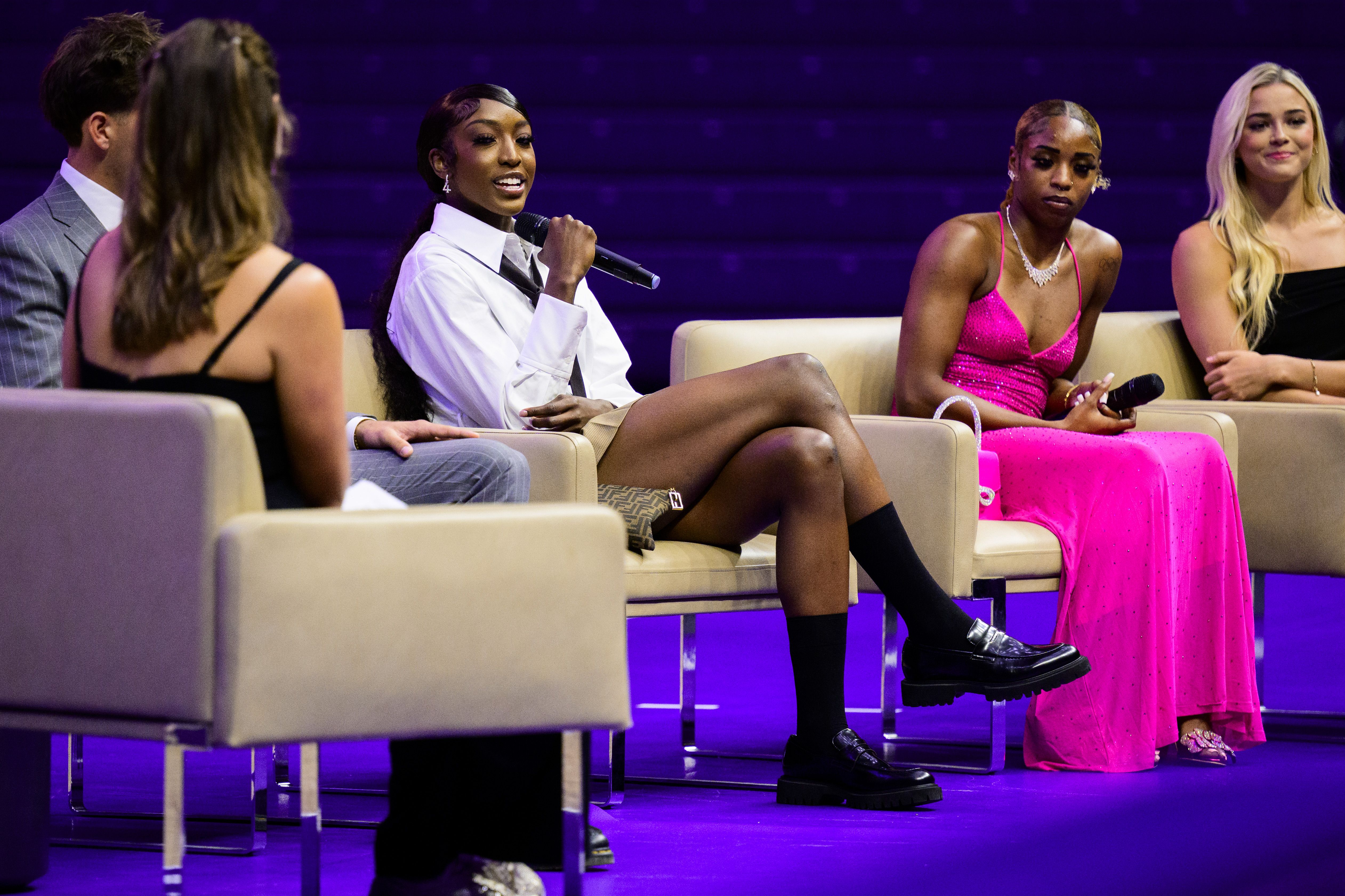 A person holds up a microphone to speak while sitting in a club chair alongside other people also sitting in chairs.