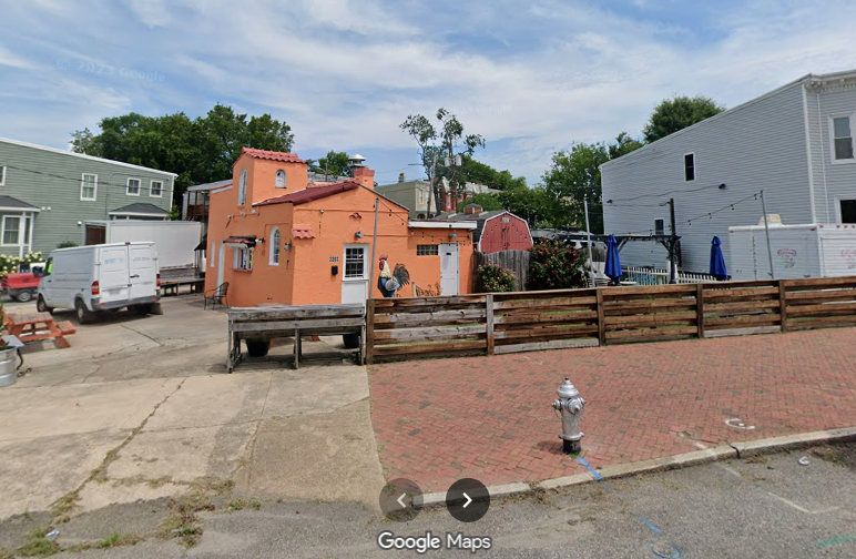 Sunlit street with a peach stucco building and rooster mural, white door, and arched windows. A wooden fence and brick plaza in front; a white van nearby, with trees and blue sky.