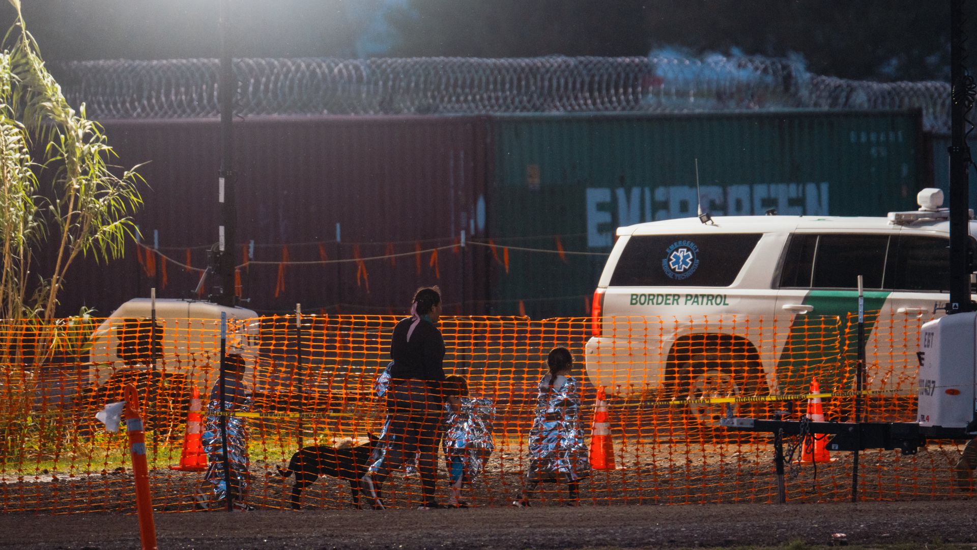 A woman and three children wrapped in mylar blankets walk past a dog and a border patrol truck at the Mexico-Texas border. 