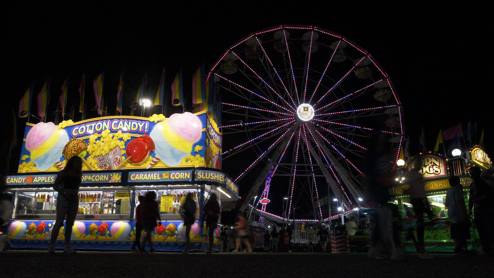 Night carnival scene with a large Ferris wheel on the right, pink and purple neon spokes; a colorful cotton candy booth on the left and silhouetted people in the foreground.