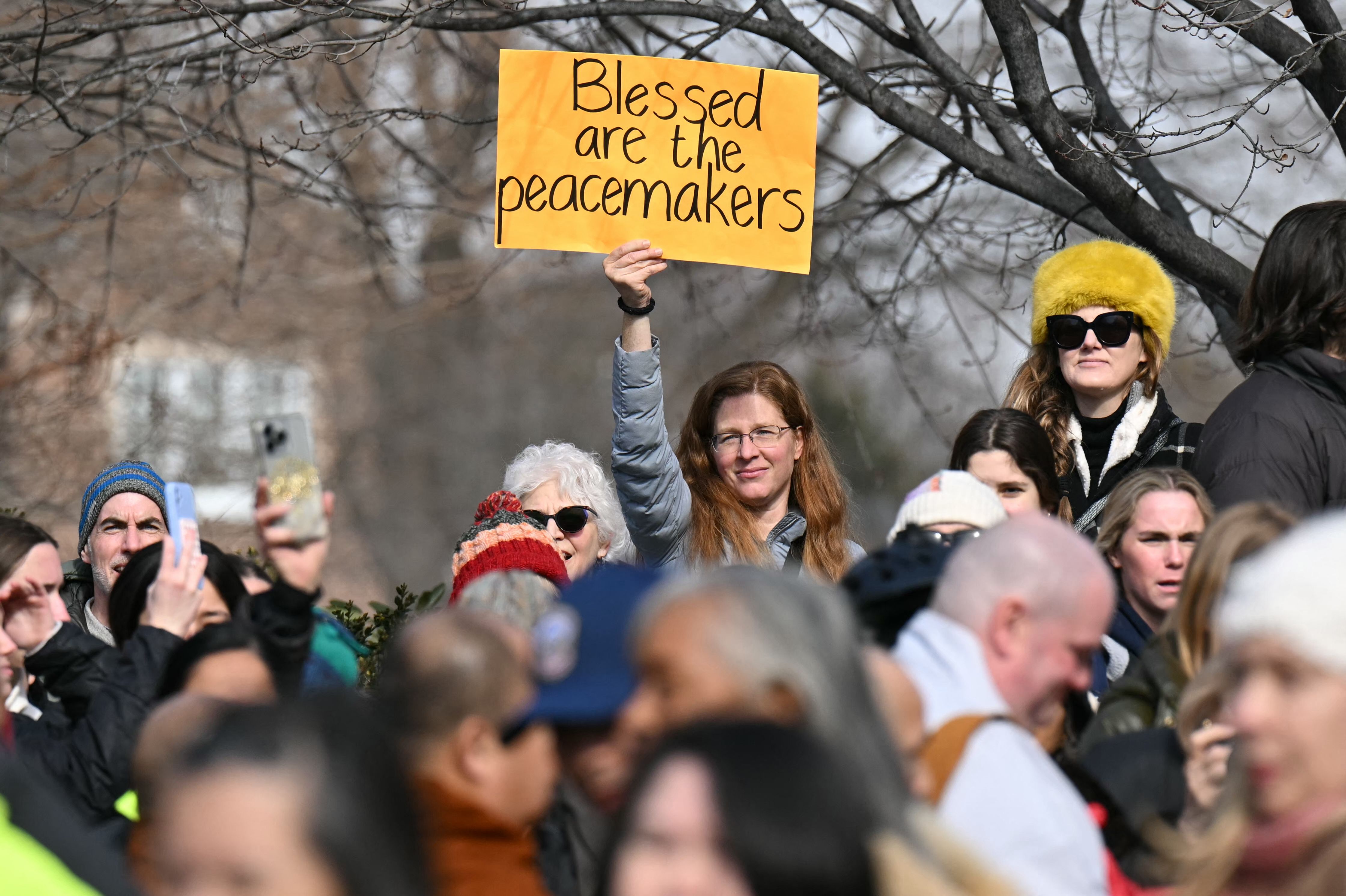 A woman in a crowd holds a bright orange sign reading "Blessed are the peacemakers". People are dressed in winter clothes, including a woman wearing a yellow fur hat and dark sunglasses.