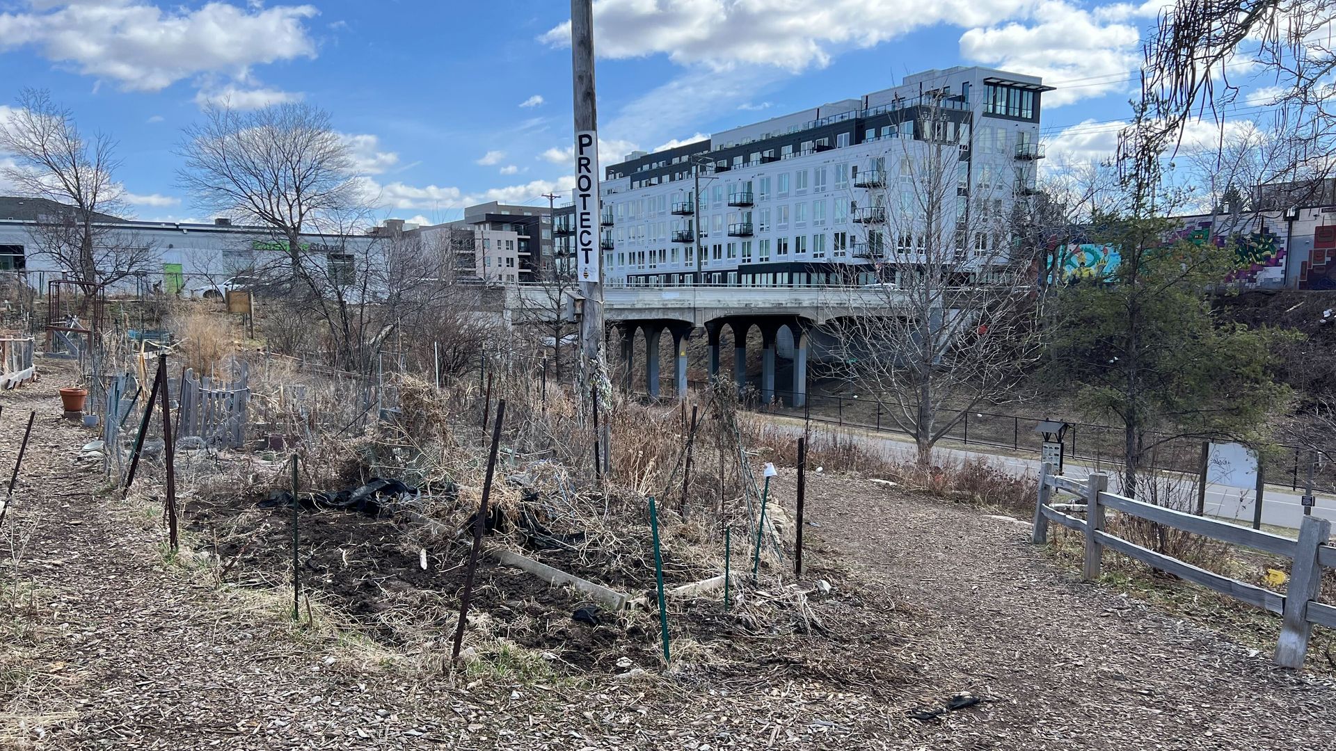 A mulch path running through a public garden with multiple plots cordoned off by chicken wire and metal posts. In the background, a bike and pedestrian path runs under an overpass and the word "Protect" appears on a utility pole.
