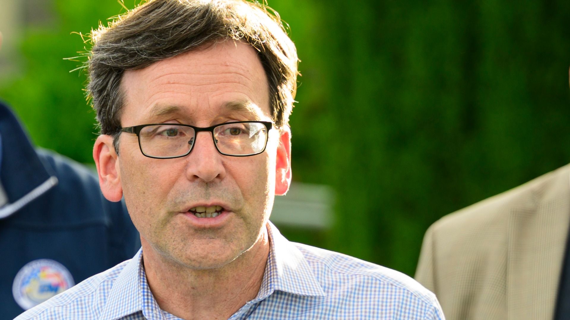 Close-up of a Bob Ferguson wearing black rectangular glasses, short brown hair, and a light blue checkered shirt, speaking outdoors with a blurred green background.