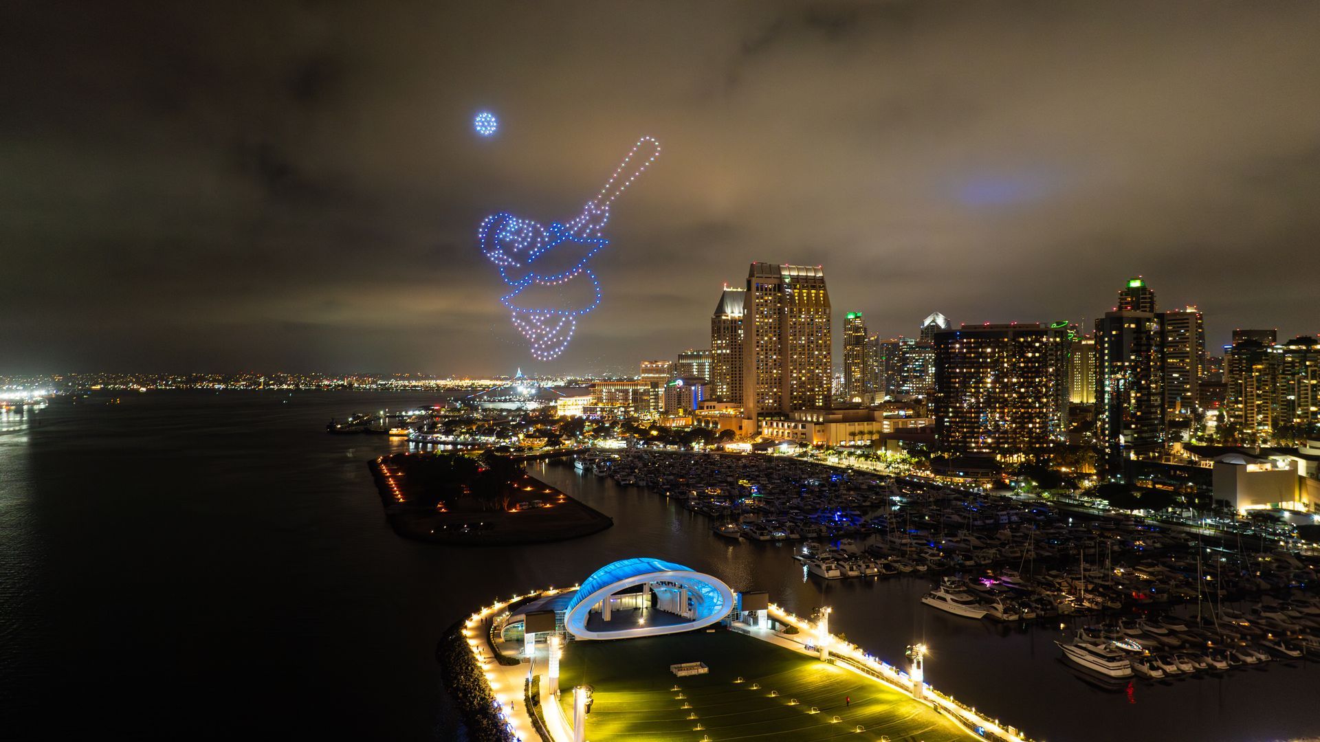 Nighttime San Diego city skyline along a harbor with boats; a large blue dotted drone light sculpture of a baseball player hitting a ball above the buildings and lit up Rady Shell music venue.