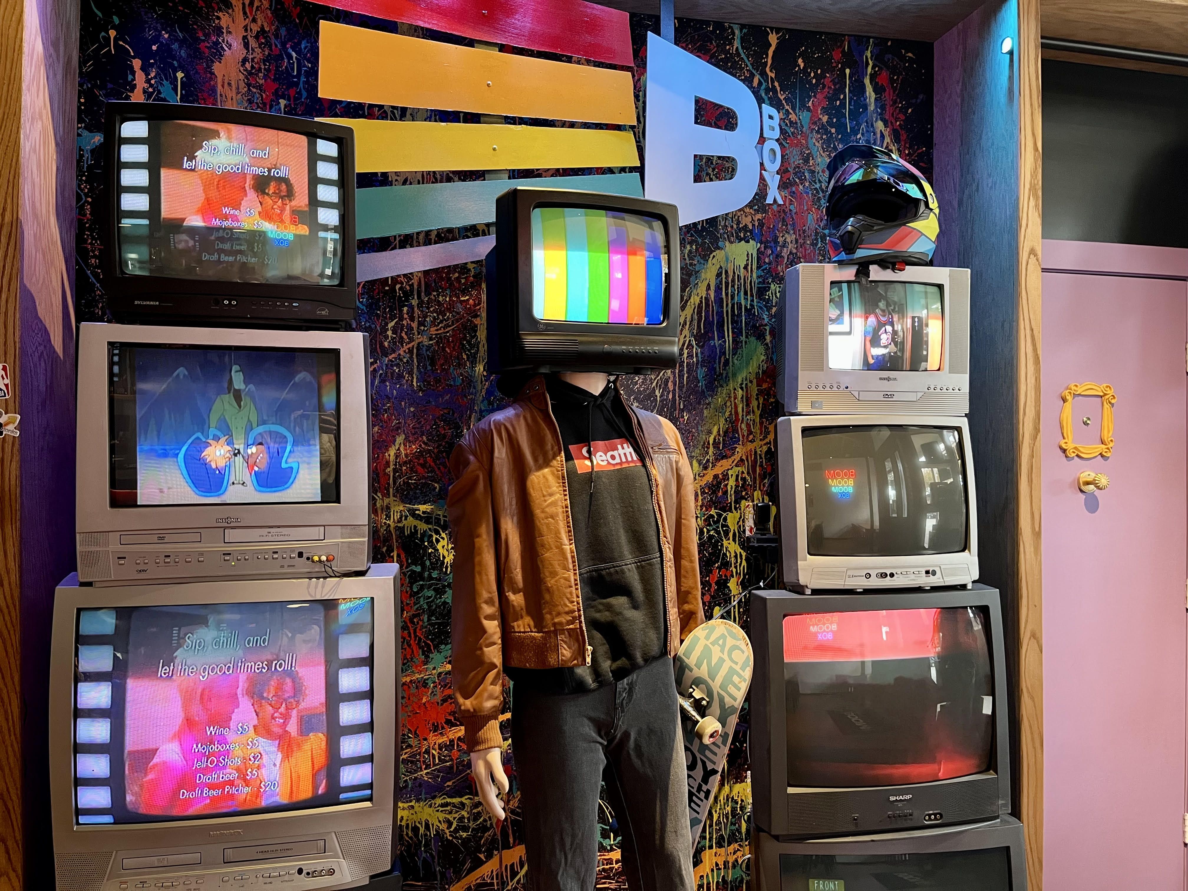 A wall full of TVs and a mannequin with a TV as a head, next to the "Friends" purple door.