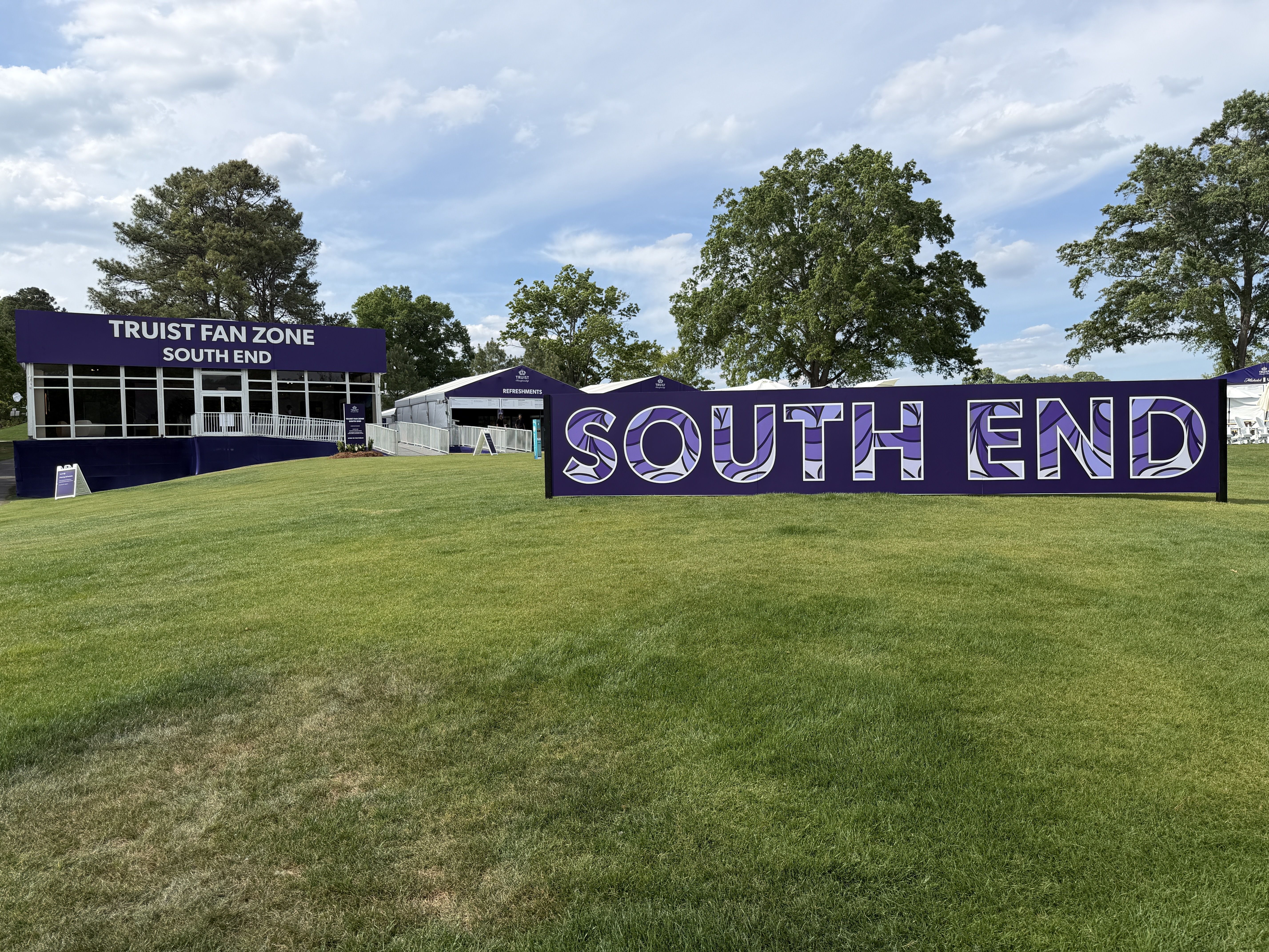 Wide grassy field with a purple event building labeled "TRUST FAN ZONE SOUTH END" and a long purple banner reading "SOUTH END"; trees and a blue sky with white clouds in the background.