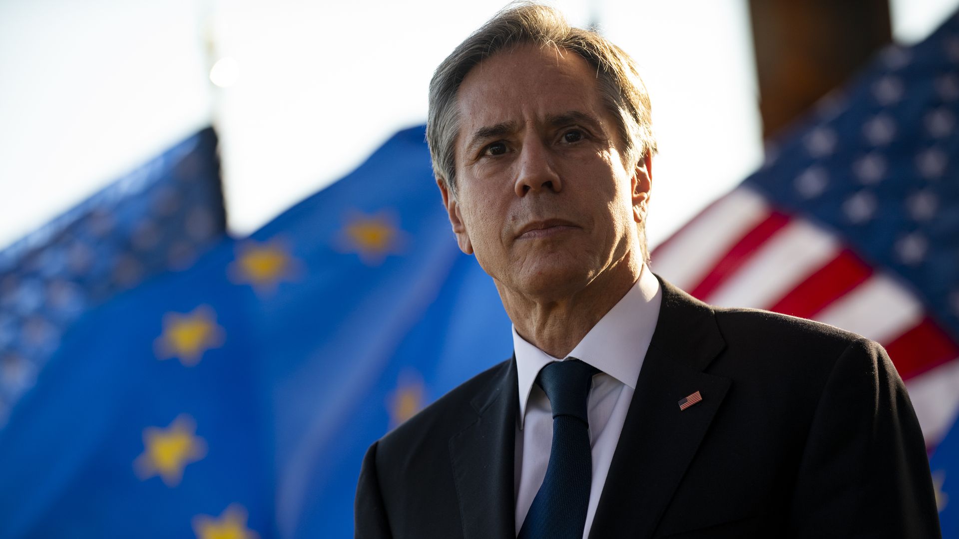 Photo of Antony Blinken in a suit standing against the backdrop of the American flag