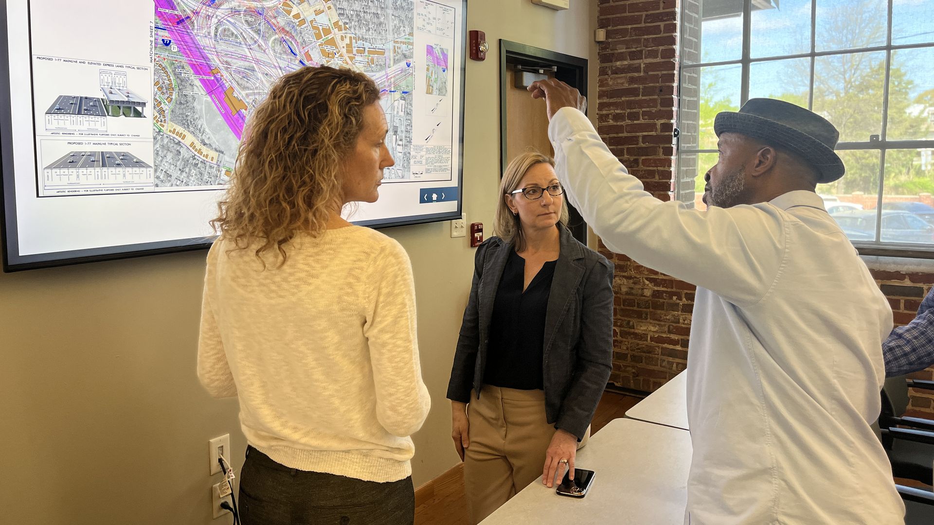 Three professionals study a large map on a wall screen in a bright room; a man in a white shirt and hat points to the map while two women, one with curly hair and a blazer, look on.