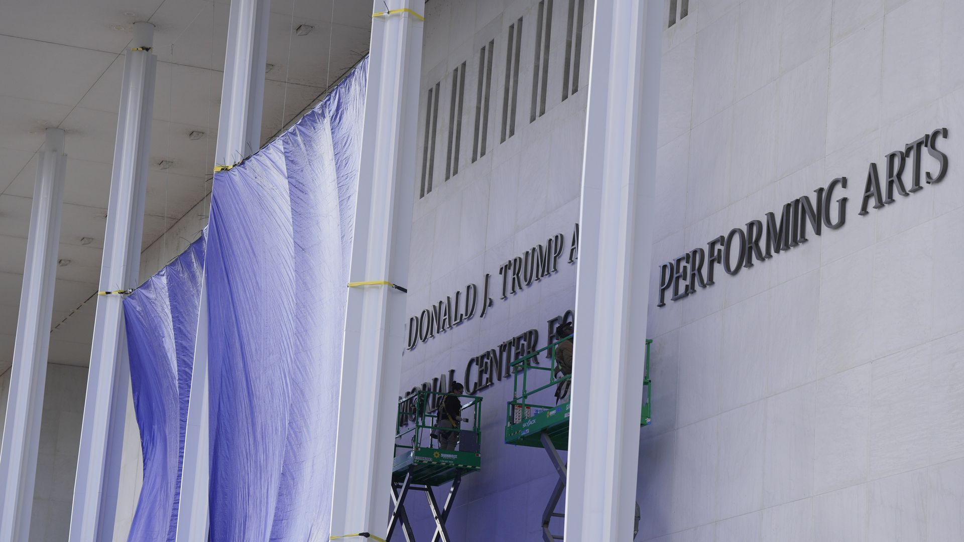 Workers in lifts working on letters on a white building with columns and a large purple tarp hanging beside them.