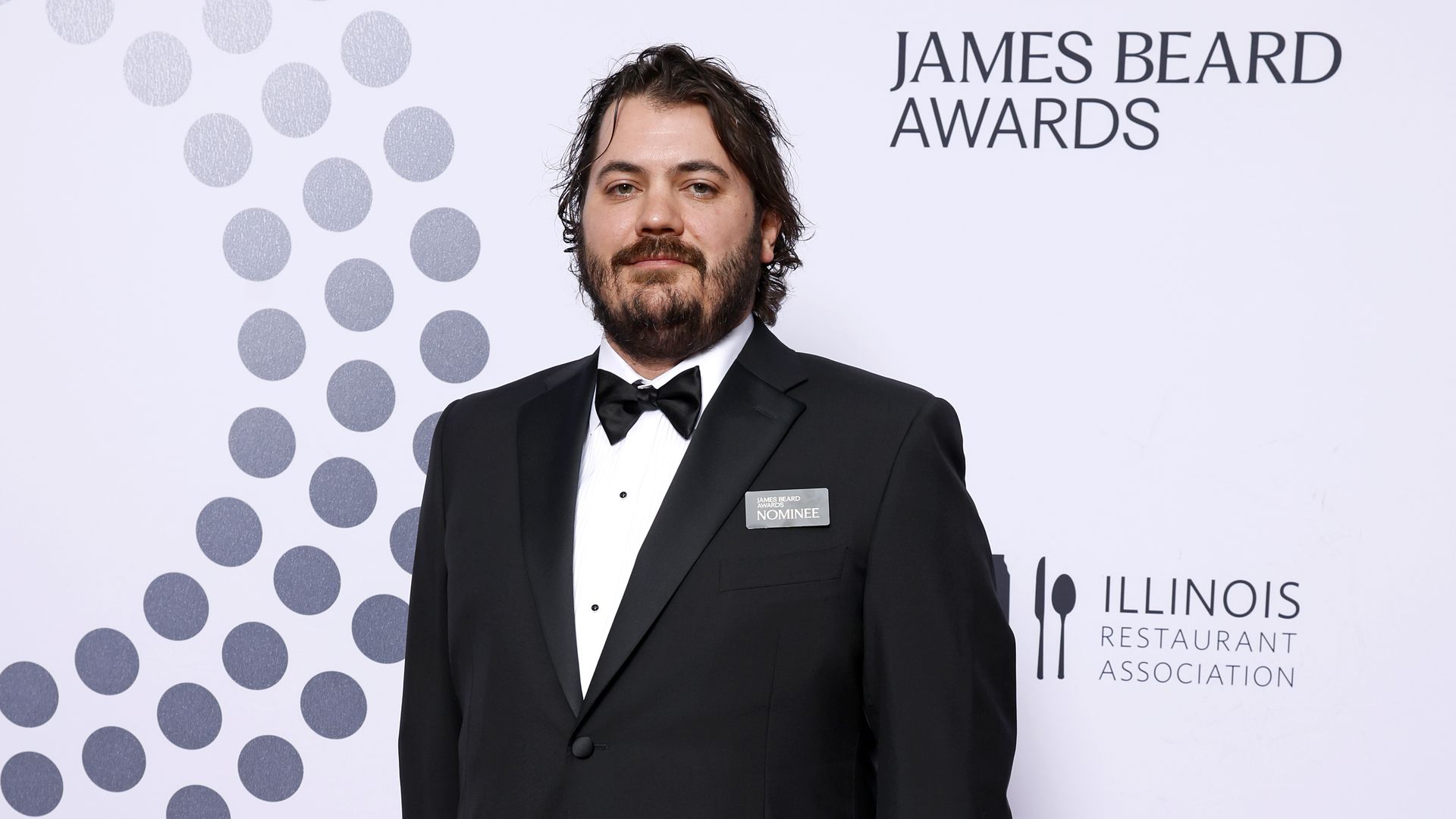 A man in a black tuxedo stands with his hands in his pockets on an awards show red carpet
