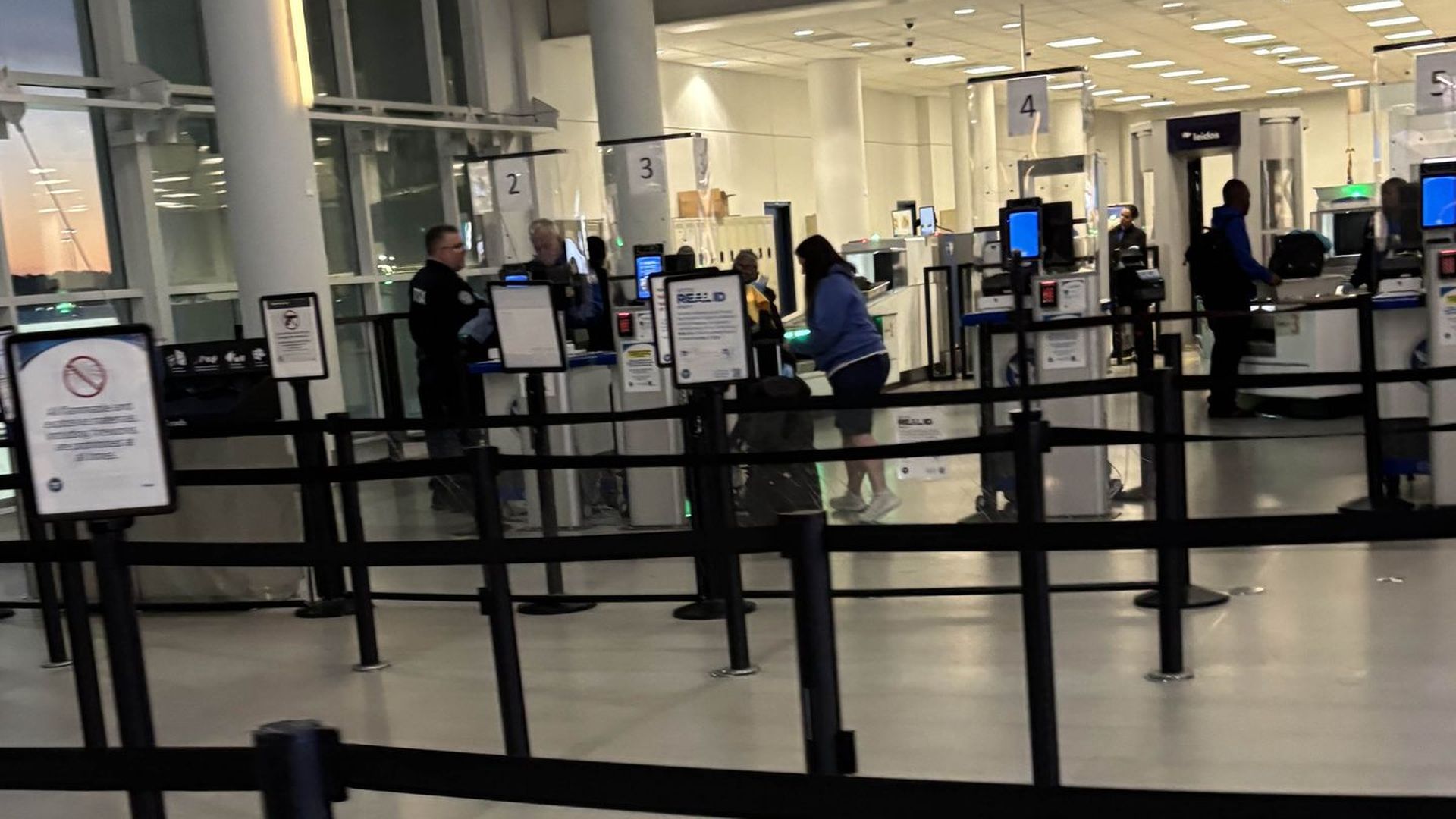 Airport security checkpoint with lanes 2–5, black belt barriers, travelers and staff at kiosks, illuminated screens, and large windows showing a sunset outside.