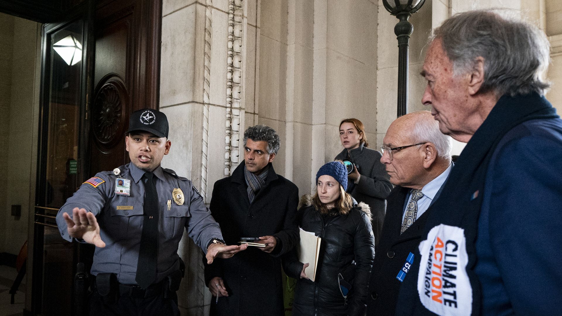 A security guard in a gray uniform gestures to a group of lawmakers and protesters outside a marble building.