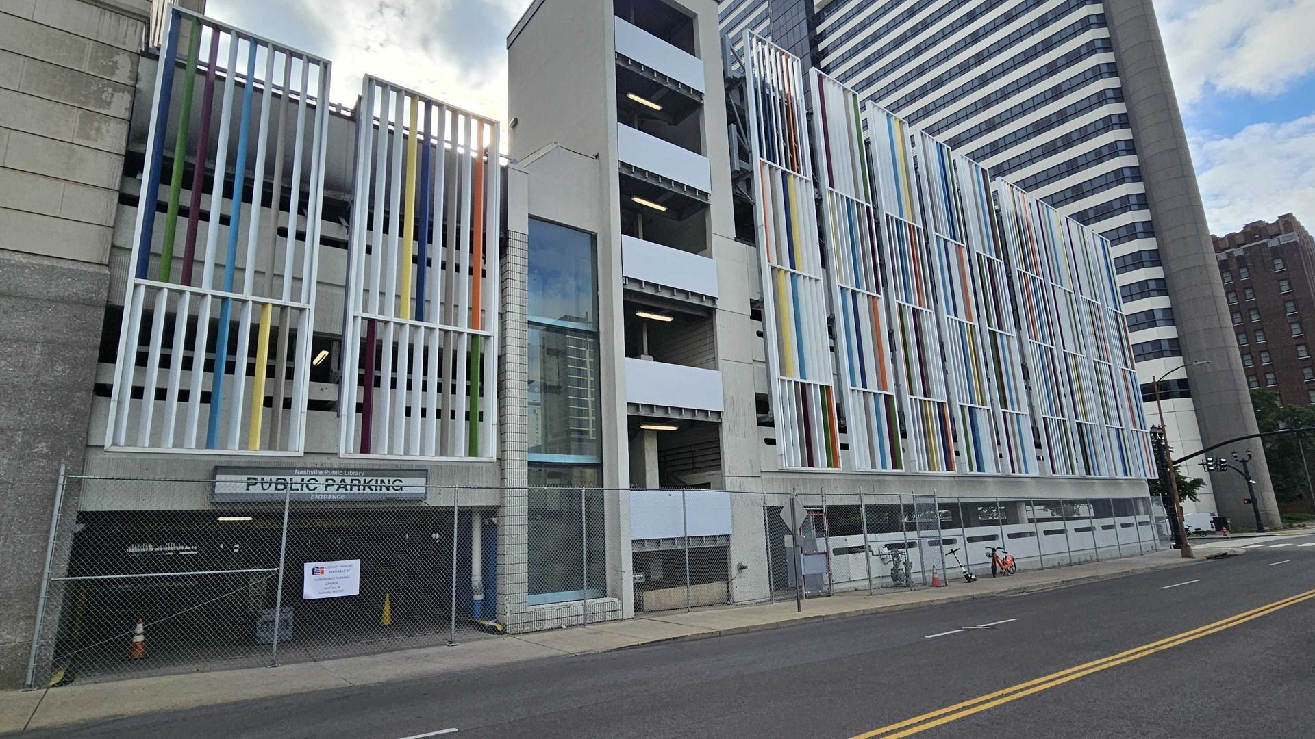 Street view of the library parking garage facade with vertical multicolor slats (blue, green, orange, yellow) in front of a modern high-rise; chain-link fence and blue sky.