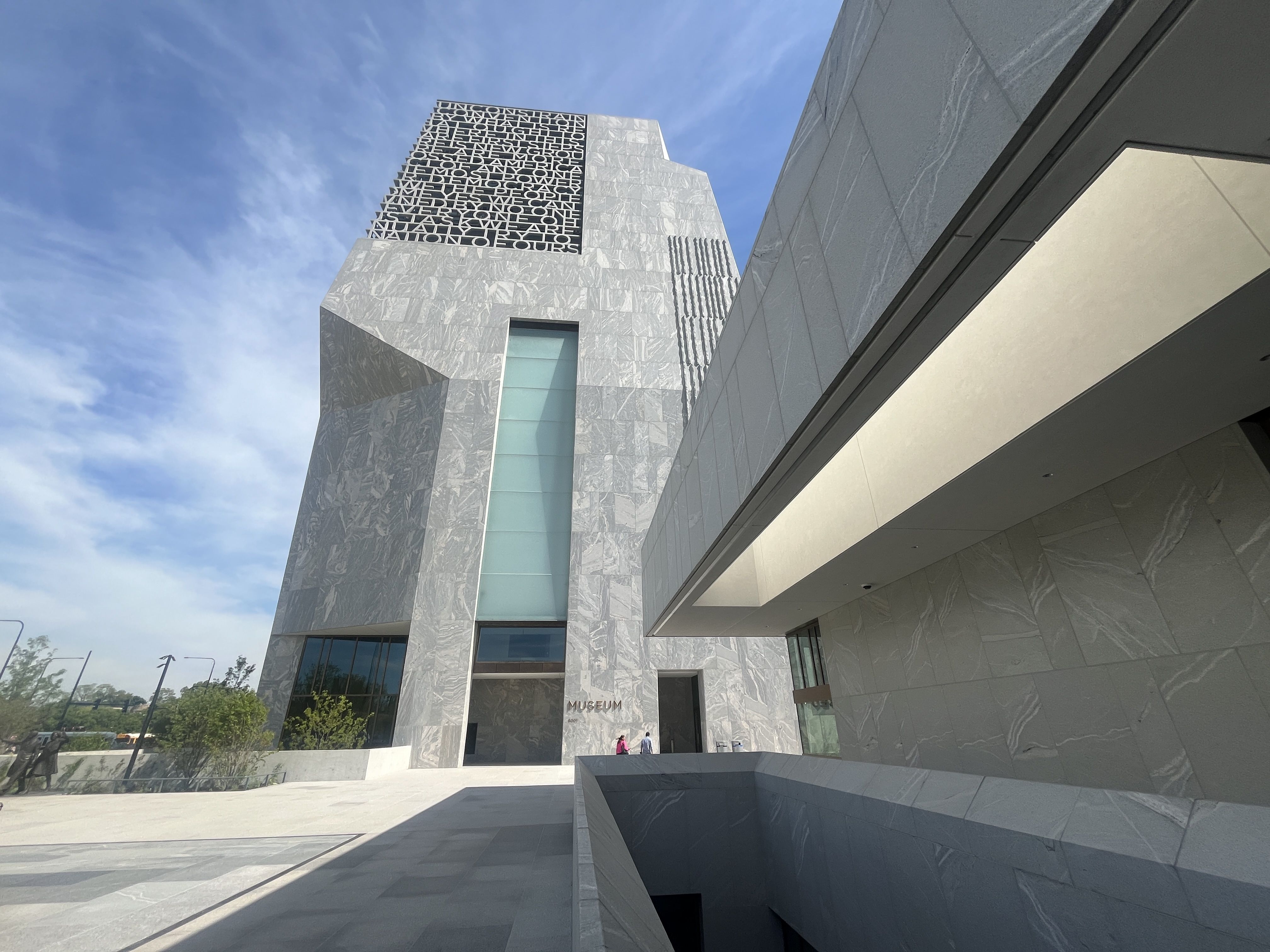 Modern silver marble museum facade with angular blocks and a large black square panel of letters. Glass windows, a covered entrance, and a few people near the doorway under a blue sky.