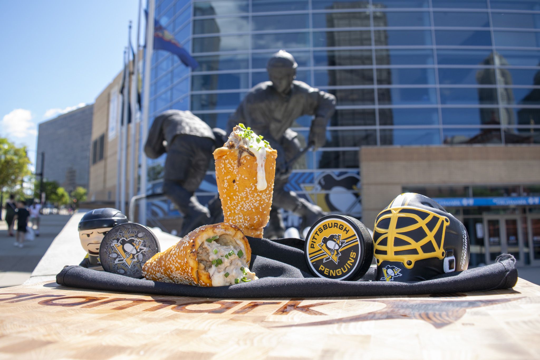Close-up of a sesame-coated sandwich with creamy sauce and chopped green onions on black fabric; blurred background shows a glass building, a statue, and Pittsburgh Penguins helmets.