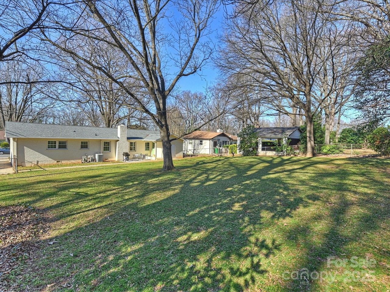 Large grassy backyard with leafless trees casting long shadows, a white brick house and two small sheds or outbuildings in the background under a clear blue sky.