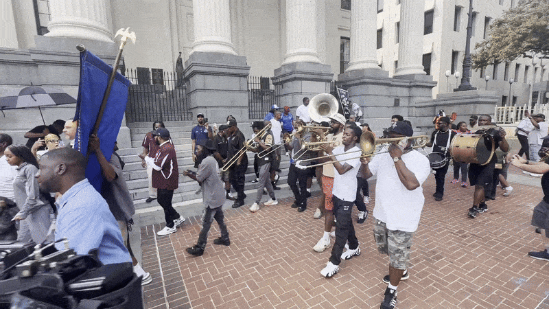 Marching band in casual clothes plays trombones, sousaphone, and drums while walking on a brick path outside a building with large columns during daytime.