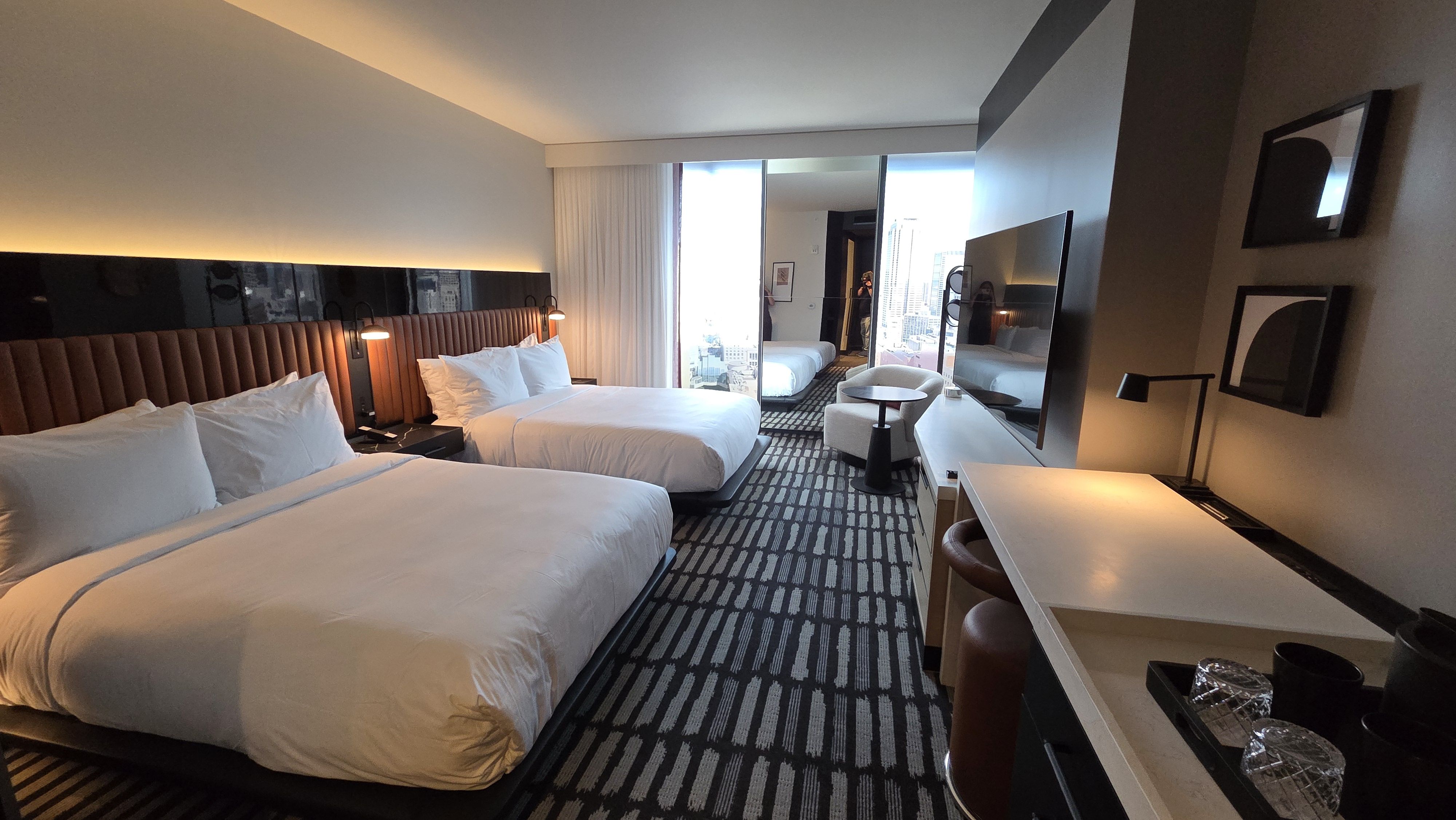 Modern hotel room with two large beds, white bedding, black and brown furniture, a wall-mounted TV, a desk with a lamp, and large mirrors reflecting city buildings outside the window.