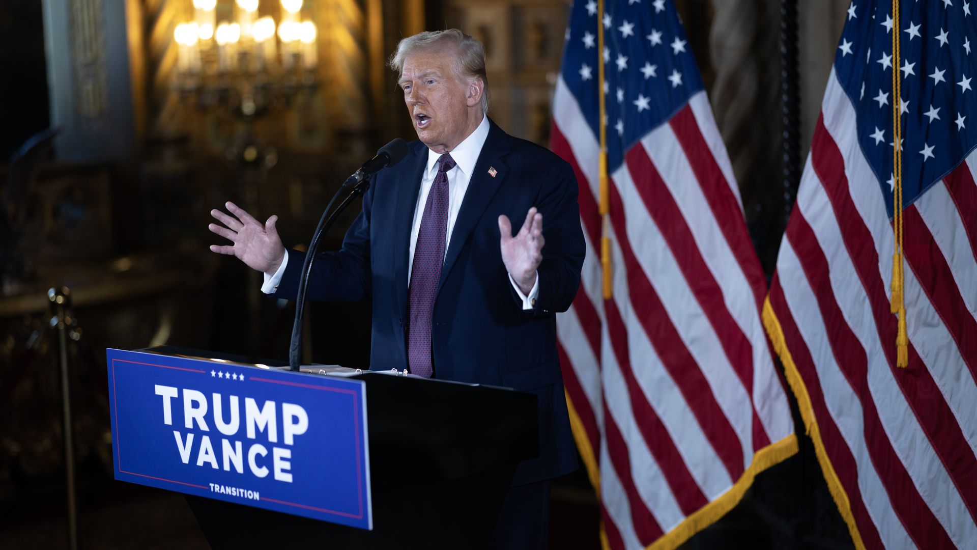 Donald Trump, wearing a blue suit and speaking at a "Trump Vance" podium in front of American flags.