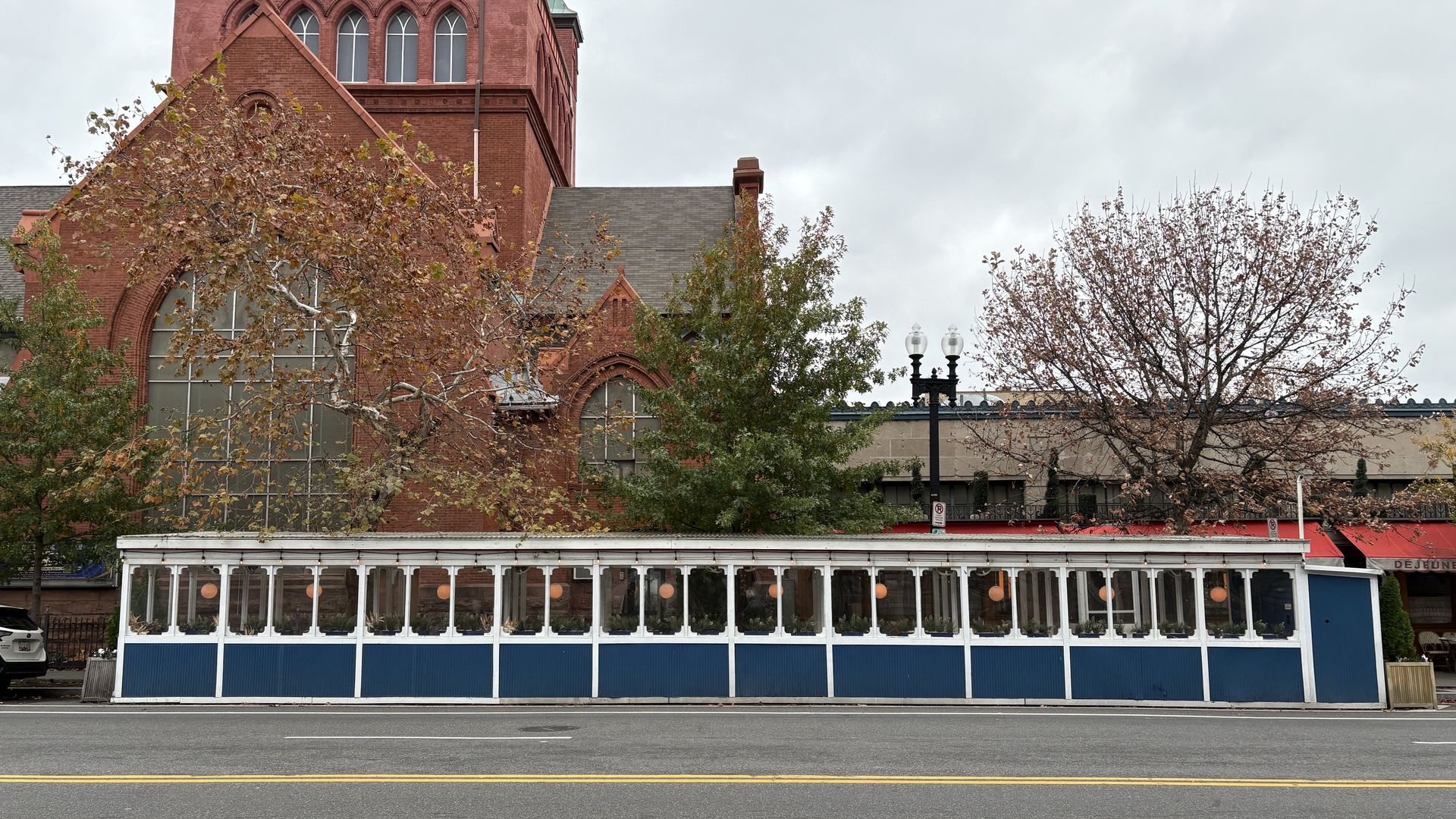 A blue and white enclosed outdoor dining area on a city street, with a red brick church and trees with autumn leaves in the background under a cloudy sky.